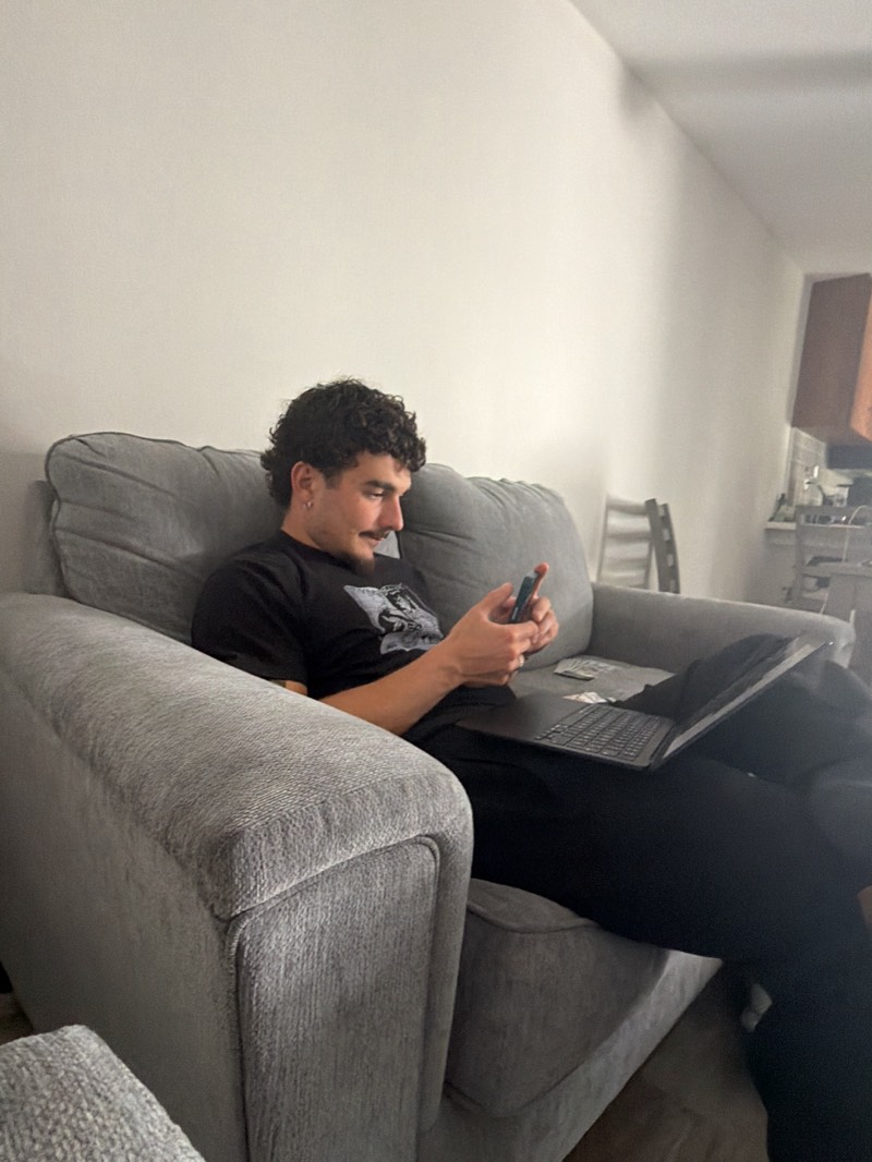 A man with dark, curly hair, a mustache, and a short beard sits comfortably on a large, grey fabric couch. He wears a black t-shirt with an indistinct graphic and black pants, along with an earring in his left ear. He holds a smartphone in both hands, looking down at its screen with a slight smile, suggesting amusement or contentment. An open black laptop rests on his lap, and some folded papers or money are visible next to it on the couch.
The grey couch features thick cushions and armrests. The background walls are plain and light-colored. To the right, a partially visible kitchen area with cabinets and appliances, and a light-colored chair or stool, suggests an open-plan living space. The casual indoor setting, likely a living room or common area, takes place in Redondo Beach, United States, depicting a moment of relaxation or casual engagement with technology. No legible text is visible in the image.