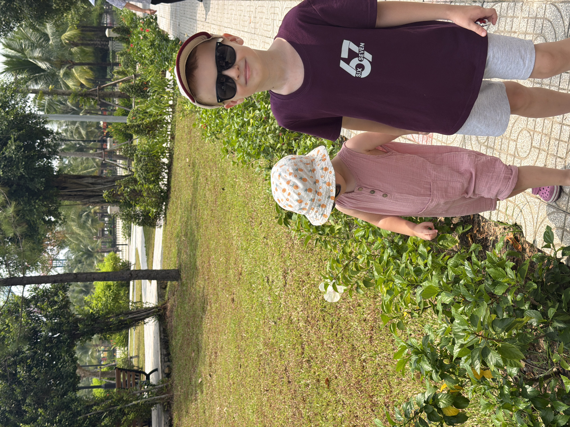 Two children, a boy and a younger child, stand on a paved walkway in a park. The boy, on the right, is wearing a maroon t-shirt with a graphic of "67 SIX SEVEN" and grey shorts. He has on sunglasses and a baseball cap. The younger child, on the left, wears a pink romper and a bucket hat with a pineapple pattern. They are both facing forward. Lush green grass and trees are visible in the background, suggesting an outdoor recreational area. The setting appears to be a sunny day in a city park, likely Ho Chi Minh City, Vietnam, given the presence of palm trees and tropical vegetation.