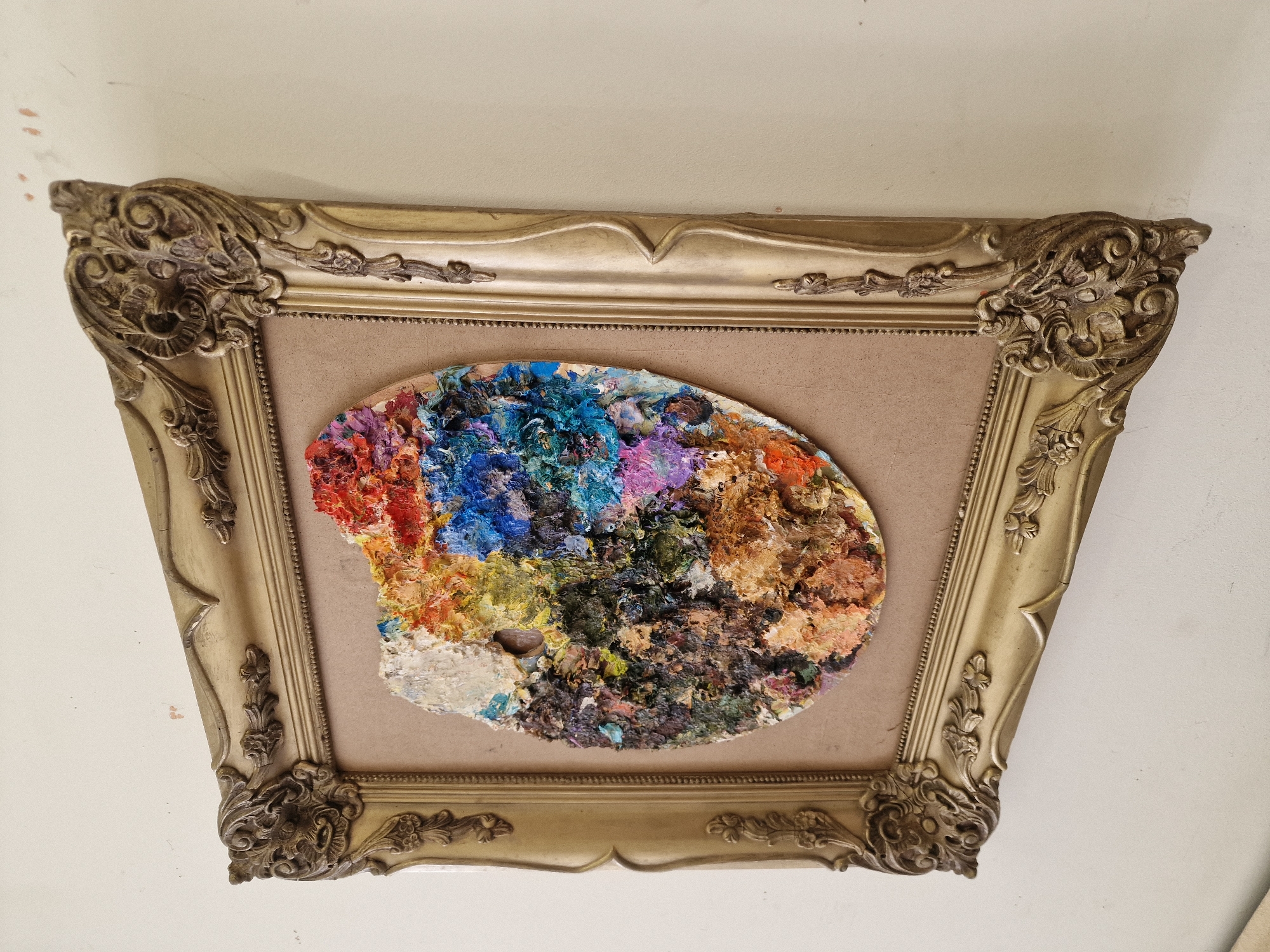 A framed artist's palette is depicted hanging on an interior wall in the city of Veliko Tarnovo, Bulgaria. The central subject is an oval-shaped wooden palette, approximately 30-40 cm in length, with a thumbhole. Its surface is densely covered in thick, dried applications of oil paint, creating significant impasto texture. A wide spectrum of colors is present, including vibrant reds, blues, yellows, greens, purples, and mixed earthy tones. This paint-laden palette is mounted on a plain, light brown, rectangular backing board. The entire piece is enclosed within a highly ornate, gilded wooden frame. The frame features intricate relief carvings, particularly at its four corners and along its inner and outer borders, consistent with classical or Baroque decorative styles. The gold finish exhibits signs of age and minor wear. The frame and its contents are displayed against a plain, off-white wall, which shows minimal surface imperfections and faint marks. The scene is illuminated by ambient light, casting soft shadows that highlight the textures of the paint and the frame's ornamentation. No individuals are present, and no actions or interactions are depicted.