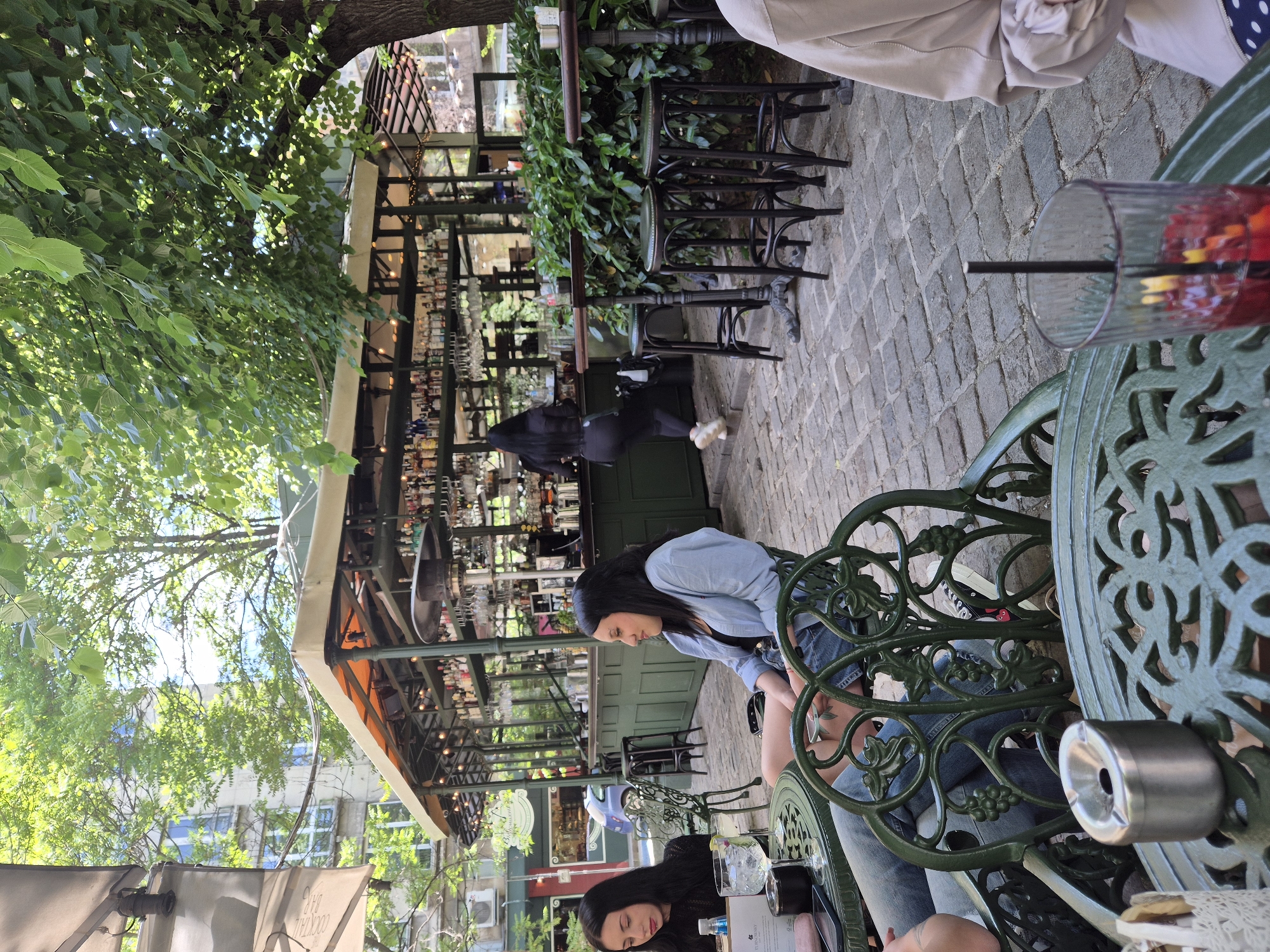 The image shows a partially outdoor cafe scene in Unknown, Unknown, with two women seated at a green wrought-iron table. The table has an ashtray on it and a cocktail glass filled with a red drink. One woman is wearing a light blue top, and the other is wearing a black top. Behind them is the bar area of the cafe, which is well-stocked with bottles. Behind the bar is a person wearing black and white shoes. The cafe has a leafy green overhead structure. The ground is cobblestone, and there is a tree with dense foliage visible in the top left corner.