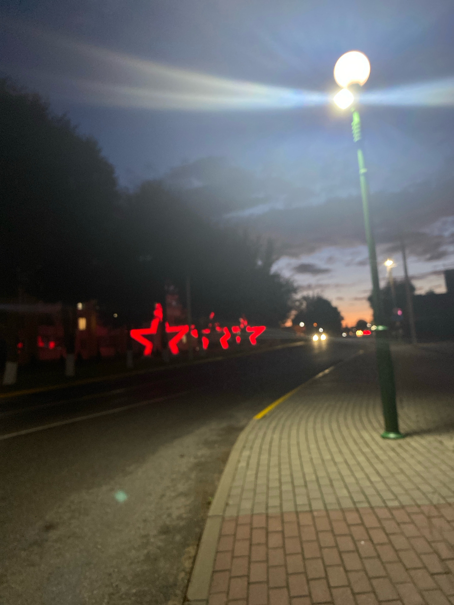 The image captures a street scene at dusk, likely in Voranava, Belarus, with a focus on decorative red star-shaped lights lining the road on the left. The sky is a gradient of dark blue and purple, with clouds visible and a bright light source, possibly a street lamp, dominating the upper right quadrant. The street itself is paved, with a sidewalk made of interlocking bricks on the right. Several vehicles are present on the road, indicated by their headlights, and some red lights that appear to be part of a decoration or sign are visible on the left. The image has a soft focus, giving it a slightly blurred, atmospheric quality.