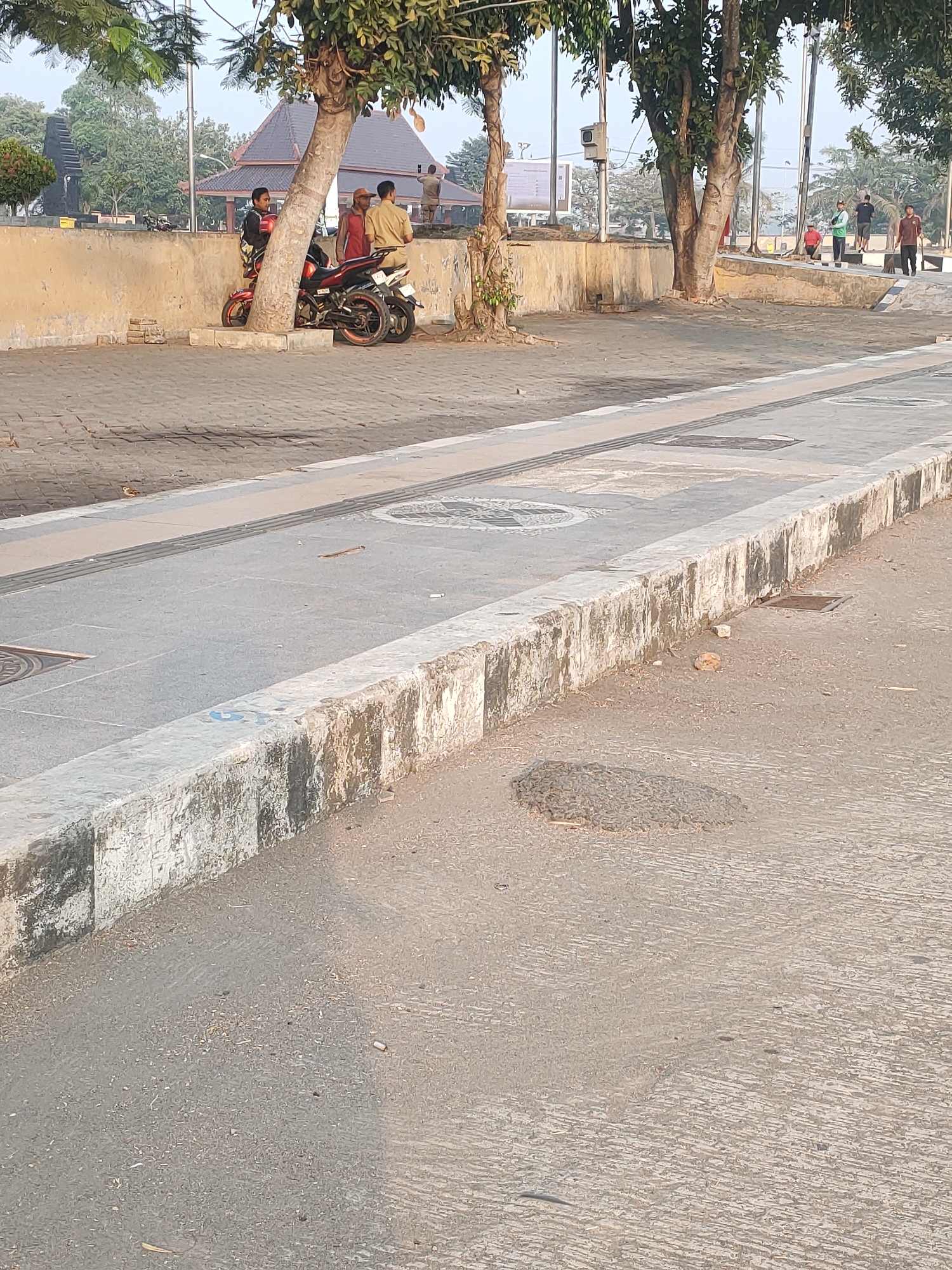 The image shows an outdoor scene in the city of Kanigoro, Indonesia. In the foreground, a grey concrete curb is visible, with dirt and moss growing on its surface. Beyond the curb is a paved area with a textured surface, possibly asphalt, with some small debris scattered across it. Further back, a paved walkway with inlaid textured strips runs parallel to the curb. In the background, several people are standing and walking. Two individuals are seated on a red motorcycle parked near a stone wall. Several trees are scattered throughout the scene, with a large tree in the mid-ground. A building with a dark, pitched roof is visible behind the wall. The sky is hazy and pale, suggesting a dry or dusty atmosphere.
