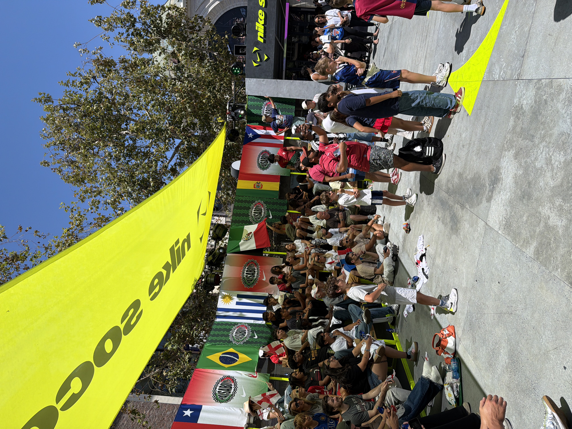 This is a wide shot of an outdoor event in Los Angeles, California. A large, bright yellow banner with black text is draped overhead, partially obscuring the sky and a leafy tree. Below the banner, a row of flags hangs, including those of Chile, Brazil, and Mexico, interspersed with other banners featuring a circular logo. A crowd of people, of various ages and ethnicities, are gathered in the scene. Many are seated on bleachers facing a raised platform, while others stand in front of it. Some individuals are holding up phones, seemingly recording the event. To the right, a "Nike Store" sign is visible, suggesting the event might be related to the brand. The ground is paved with concrete and marked with yellow arrows, indicating a pathway or designated area. Scattered on the ground are various personal belongings, including bags, water bottles, and clothing. The overall atmosphere appears to be one of a public gathering or festival.
