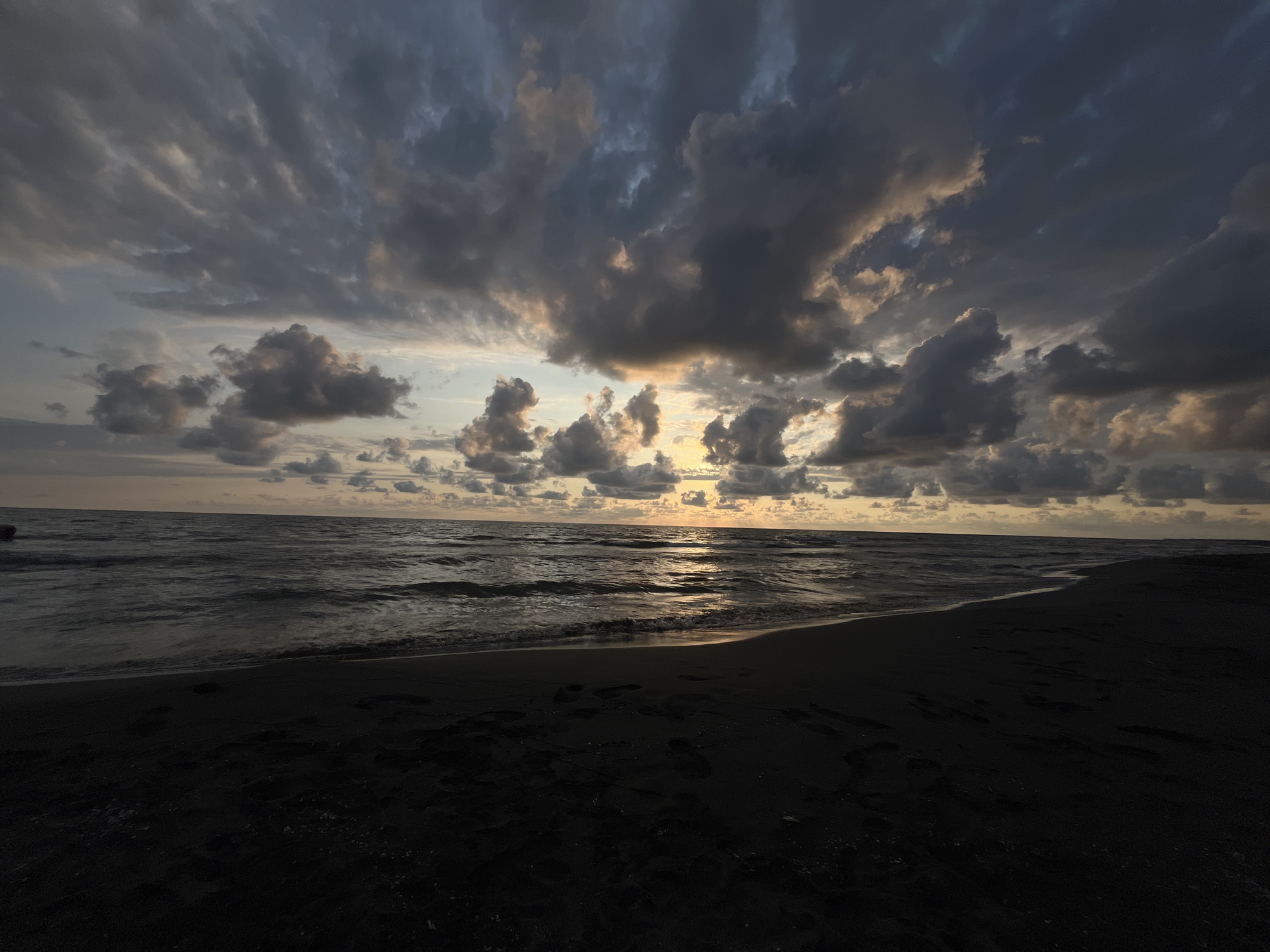This image captures a sunset over the Black Sea at the Supse Community beach in Georgia. The sky is filled with dramatic, dark clouds, with breaks of light near the horizon where the sun is setting, casting a golden glow on the water and some of the clouds. The sea is moderately rough, with small waves rolling onto the shore. The beach is composed of dark sand, with visible footprints scattered across its surface. The overall scene is moody and atmospheric, with the dark sand and stormy clouds contrasting with the warm sunset light.