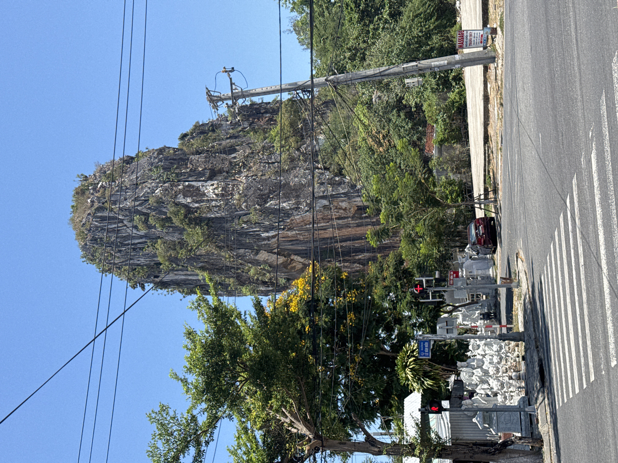 The image shows a street scene in Đà Nẵng, Vietnam. A large rock formation rises prominently behind a street cluttered with utility poles and wires. A red traffic light is visible, along with a pedestrian crossing and a row of stone statues lining the sidewalk. A maroon car is parked on the side of the road. A sign advertising "massage" is affixed to a utility pole. The sky is blue, and the overall environment suggests a sunny day.