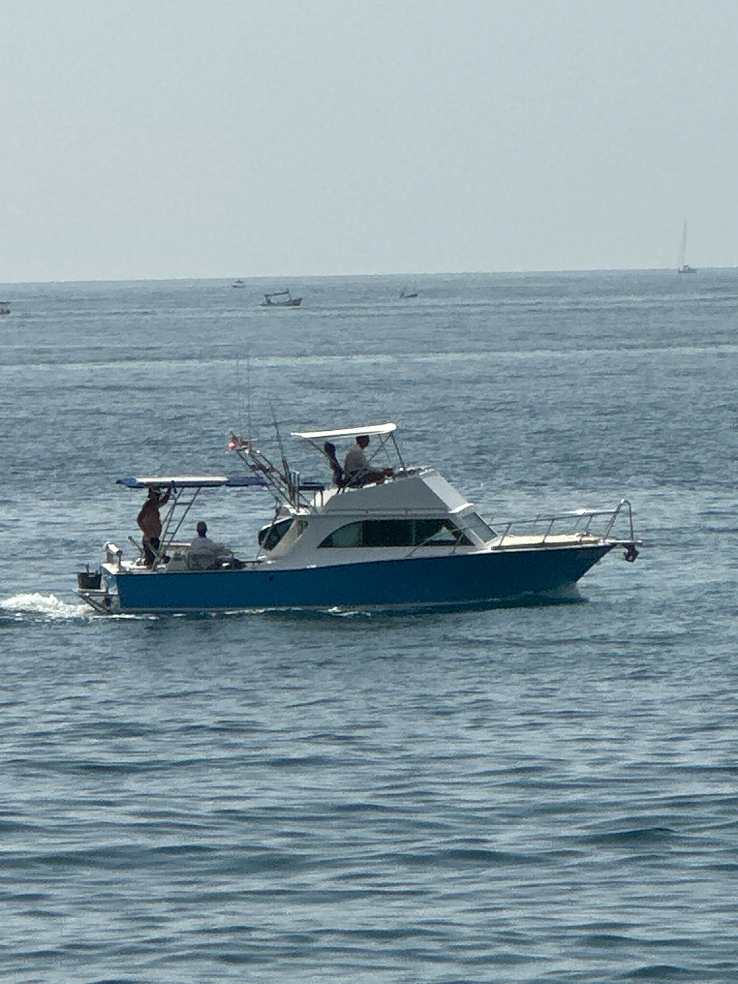 A medium shot captures a blue and white motor yacht sailing on calm, blue water under a hazy sky. The yacht has a white cabin with dark windows and a white canopy. Three people are visible on board. One person stands on the rear deck, another is seated at the helm, and a third person is visible inside the cabin through the windows. The boat is creating a white wake. In the background, the horizon is visible with several other small boats and a distant sailboat. The scene appears to be on the Adriatic Sea, likely off the coast of Budva, Montenegro, given the typical coastal scenery and boat activity in that region.
