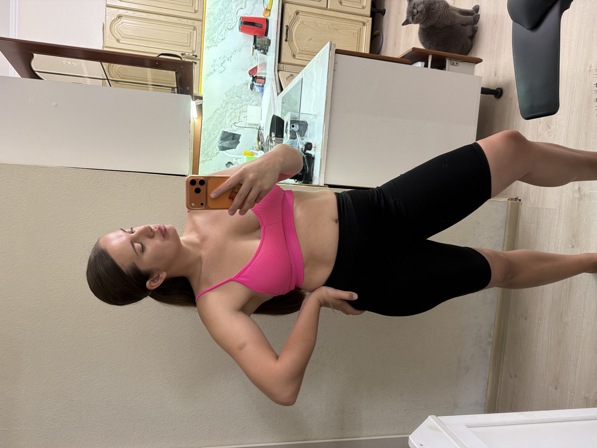 This image captures an adult female taking a mirror selfie inside a residential kitchen in Sochi, Russia. The woman, with brown hair tied in a ponytail, is clad in a bright pink ribbed sports bra with thin straps and black mid-thigh length athletic shorts. She holds an orange-cased smartphone in her right hand, angled towards the mirror, while her left hand rests on her hip. Her head is turned to her right, presenting a profile view.
The kitchen features light-colored, textured wallpaper on the left wall and light wooden laminate flooring. Behind the woman, light-toned kitchen cabinets with ornate carvings and dark handles are visible, some featuring glass panels. A light-colored countertop extends across the background, topped by a patterned backsplash and illuminated by under-cabinet lighting. Visible items on the countertop include a red electric kettle, various bottles and containers, a frying pan on a stovetop, a white sink with a faucet, and a clear plastic box. A grey towel hangs above the sink area.
In the lower right portion of the background, a grey cat sits on the wooden floor, looking towards the camera. A dark chair or stool with a black cushion is partially visible in the immediate foreground, bottom right.