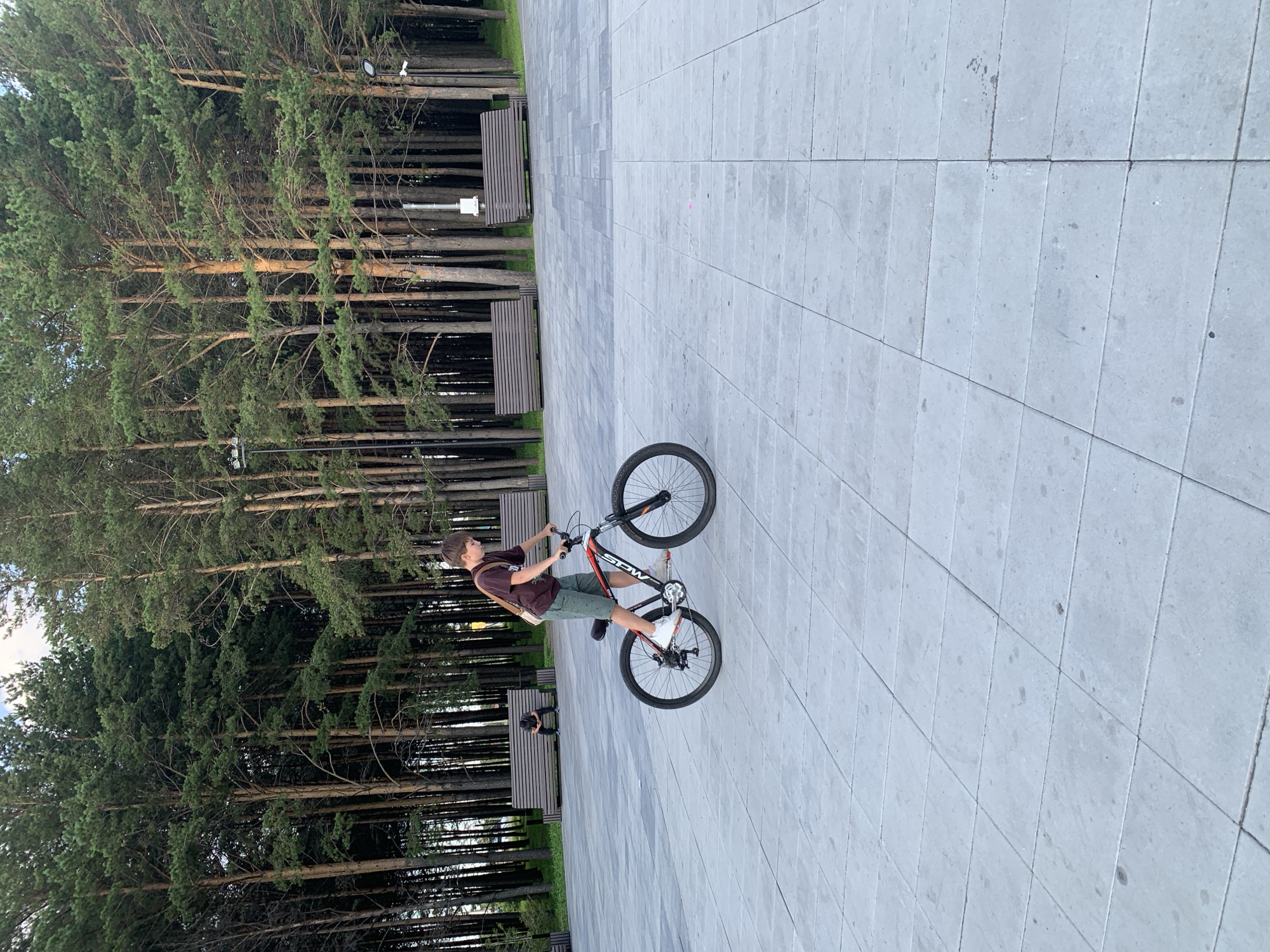 A young male is riding a bicycle on a paved area with a line of pine trees in the background. The male is wearing a maroon t-shirt, green shorts, and white sneakers. He has a brown bag slung over his right shoulder. The bicycle is black and orange with red accents. In the background, there are several wooden benches and lampposts. Two other individuals are seated on a bench in the distance. The pavement is made of large, square grey tiles. The overall setting appears to be a park or public square in Prokopyevsk, Russia.