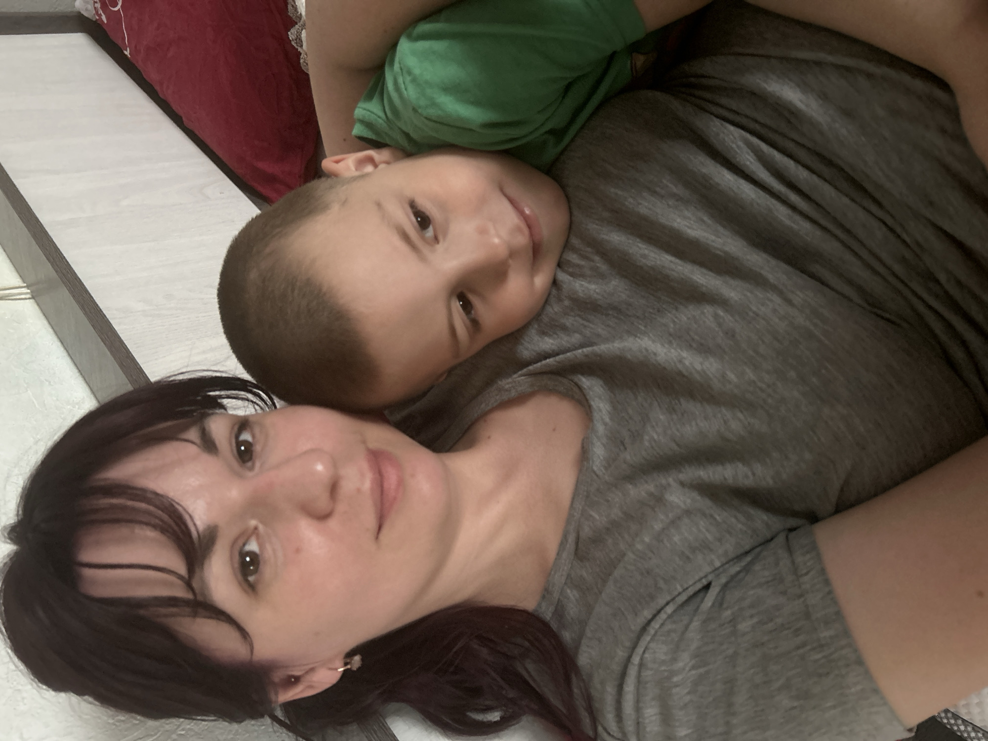 A vertical, close-up, indoor shot shows a woman with dark hair and a young boy with short, light brown hair, lying side-by-side, both looking towards the camera. The woman is in the foreground on the left, wearing a grey t-shirt and small, golden hoop earrings. The boy is in the background, resting his head on the woman's shoulder or chest, also wearing a t-shirt, possibly green. His face shows a slight smile. The woman's expression is neutral. Both subjects appear to be relaxed. The background is simple, with what looks like a wooden bed frame or floorboard on the left and a red pillow or blanket on the top right. The lighting is soft and even, suggesting an indoor setting. There is no visible text. The location is an indoor space in Foki, Russia.