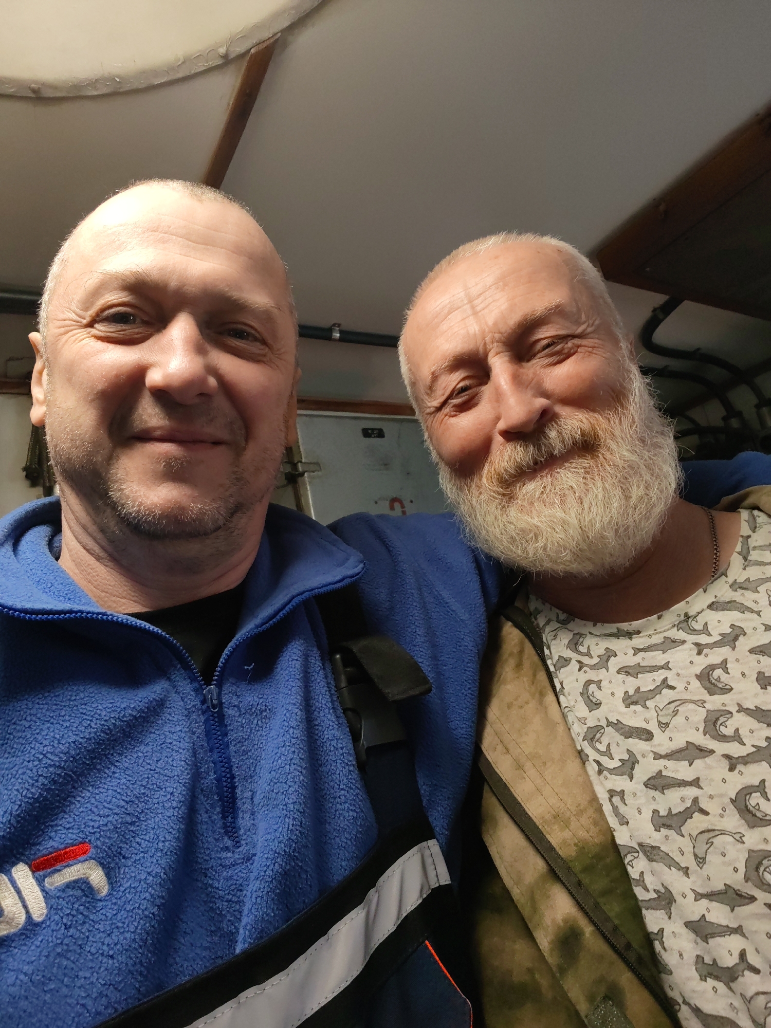 Two men are captured in a close-up, friendly selfie, likely taken indoors. The man on the left, closer to the camera, has a shaved head or very short light-colored hair and light stubble. He is smiling warmly, with wrinkles visible around his eyes. He wears a blue fleece jacket with a partial zipper, under which a dark shirt is visible. The jacket has a white and red "FILA" logo embroidered on the left chest. A dark strap crosses his left shoulder.
The man on the right, positioned slightly behind the first, also smiles, his eyes crinkling at the corners. He has a receding hairline with short, light-colored hair and a prominent, full white-to-grey beard and mustache. He is wearing a light-colored t-shirt patterned with numerous grey sharks or fish, over which he has an open, earth-toned or camouflage-patterned jacket or vest. A silver-colored chain is visible around his neck. His left arm appears to be around the shoulder of the man on the left, indicating companionship.
The background suggests an enclosed, utilitarian space with a low ceiling. The ceiling is light-colored and shows some texture or wear, with a wooden beam running across it. Visible pipes or conduits, along with a metal clamp, are present in the upper right. Part of what appears to be a metal door or hatch mechanism is visible in the middle background. The lighting is artificial, illuminating both faces. Both men convey a sense of contentment and good spirits.