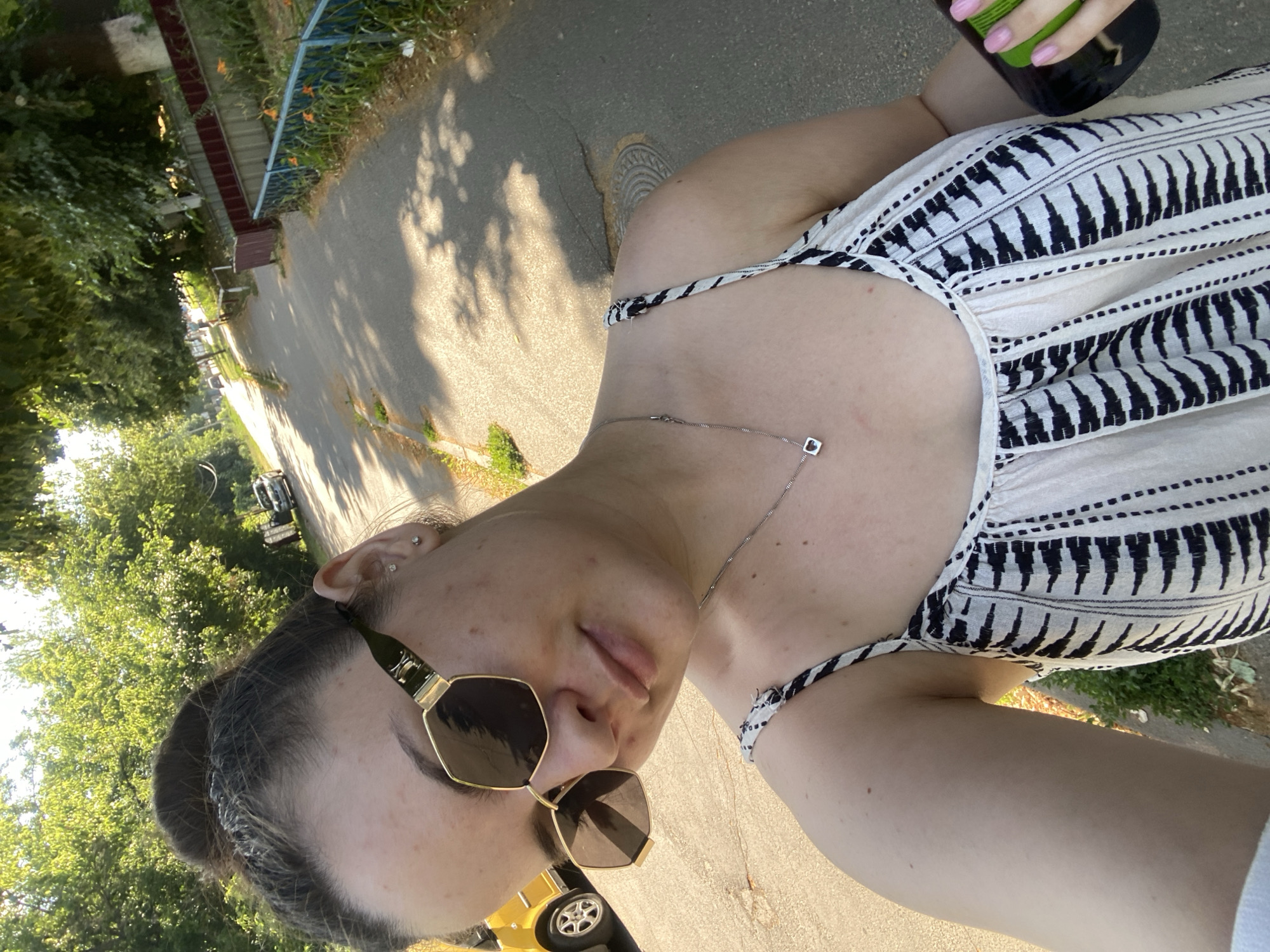 The image shows a person's upper body and face as they take a selfie. They are wearing a white and black striped dress and a necklace with a small square pendant. Sunglasses with gold frames are on their face. Their right hand, with pink nail polish, is holding a green bottle. The scene appears to be outdoors on a paved path next to greenery and a fence. Sunlight casts shadows on the ground, suggesting it's daytime. The image is oriented sideways. A yellow toy car is visible in the lower left corner. The provided location context is Poltava, Ukraine.