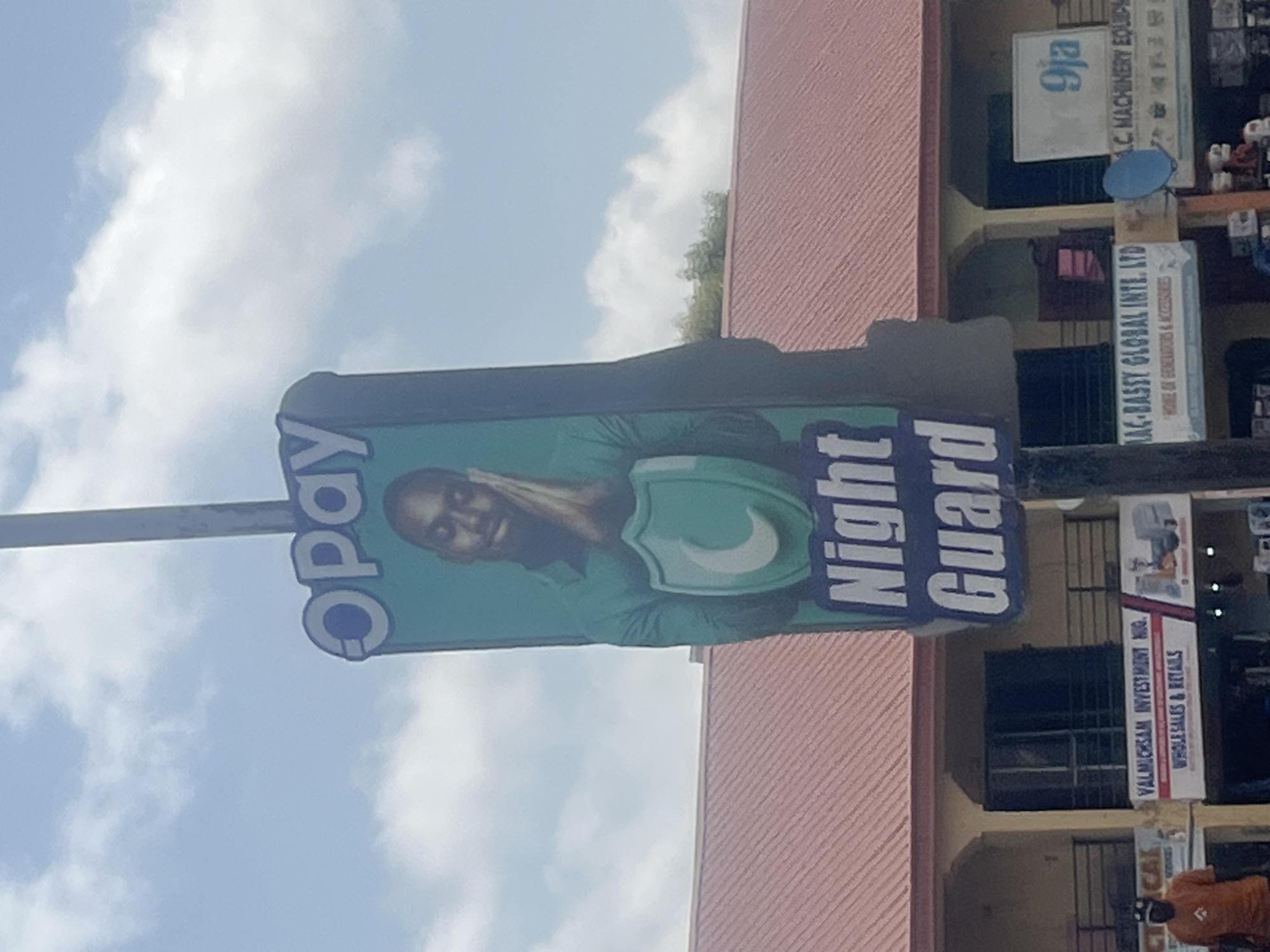 A vertical outdoor advertising sign is the main subject of this image. The sign is for "Opay Night Guard" and features a man with his hand on his head, appearing distressed, against a green background with a shield logo. The text "Opay" is on the left, and "Night Guard" is on the right of the sign, both in large blue letters.
Behind the main sign, the façade of a building with a reddish-pink striped roof is visible. To the right of the main sign, several other smaller signs are mounted on the building. One sign reads "VALMICHSAM INVESTMENT NIG. WHOLESALES & RETAILS". Another sign says "MAC-BASSY GLOBAL INTE. LTD HOME OF GENERATORS & ACCESSORIES". A smaller sign above it reads "9ja A.C. MACHINERY EQUIP".
The scene appears to be outdoors, with a bright blue sky and scattered white clouds suggesting daytime with clear weather. The angle of the photograph suggests it was taken from ground level, looking up at the signs and the building. A person in an orange shirt is partially visible at the bottom right of the frame. The overall impression is that of a commercial street scene in Nigeria, likely during daylight hours.