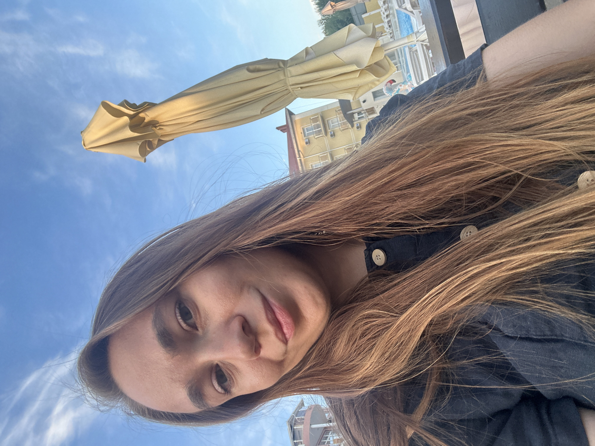 A young woman with long, light brown hair takes a selfie outdoors. She is wearing a dark blue top with buttons. Behind her, a closed, light yellow beach umbrella stands against a clear blue sky with wispy white clouds. In the background, there is a yellow building with balconies and air conditioning units, as well as a swimming pool area with lounge chairs and other closed umbrellas. The setting appears to be a resort or hotel in Vityazevo, Russia.