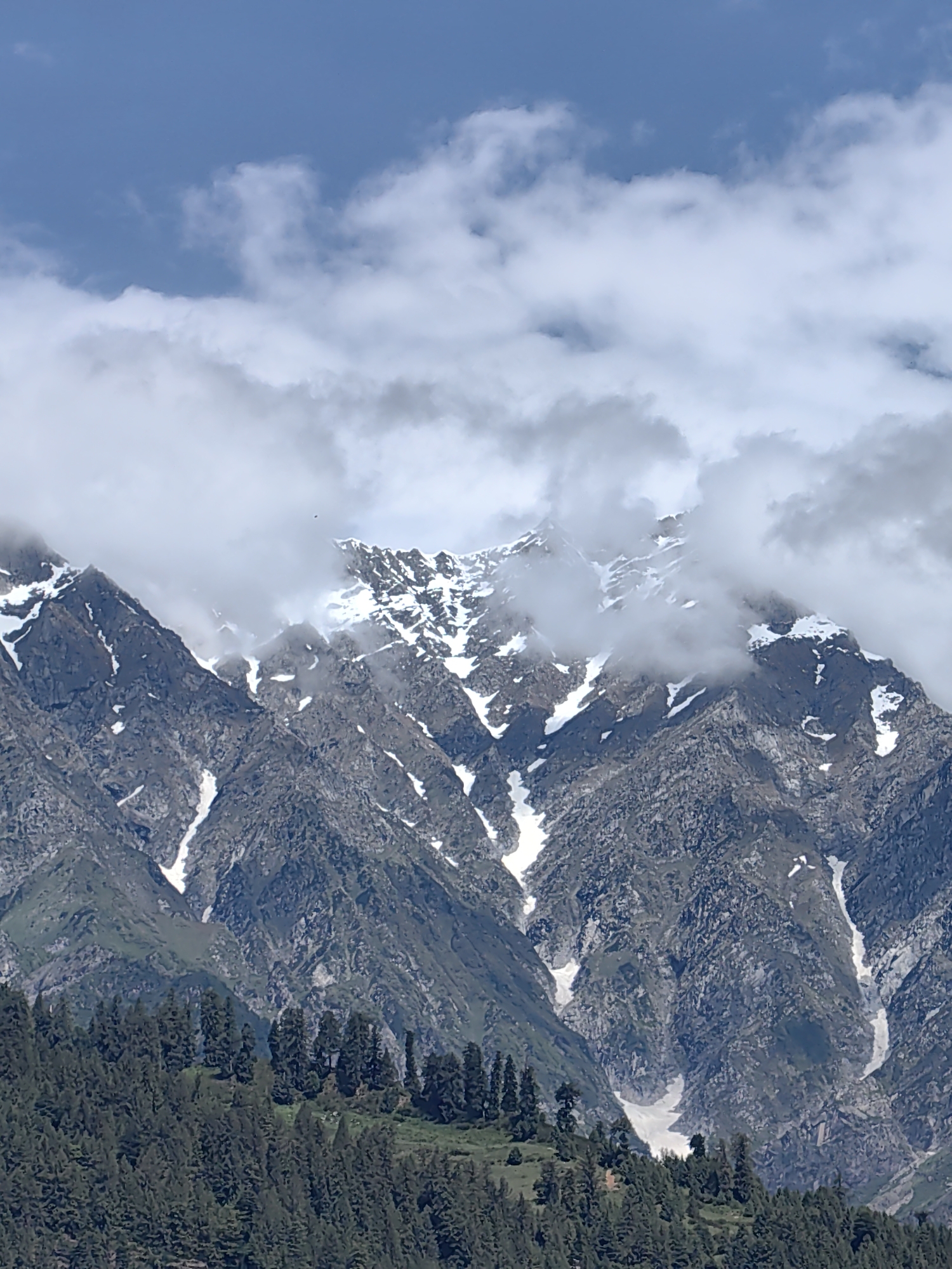 The image depicts a mountainous landscape with snow-capped peaks partially obscured by clouds. In the foreground, a dense forest of coniferous trees covers the lower slopes of the mountains. The mountains themselves are rugged and rocky, with patches of snow visible in the crevices and on the upper reaches. The sky is partly cloudy, with white clouds contrasting against the blue sky. The scene appears to be a natural landscape, likely in a region with significant elevation. Given the mention of Vashisht, India, this is likely a view of the Himalayas or a similar mountain range in that region. There are no people or animals visible in the image.