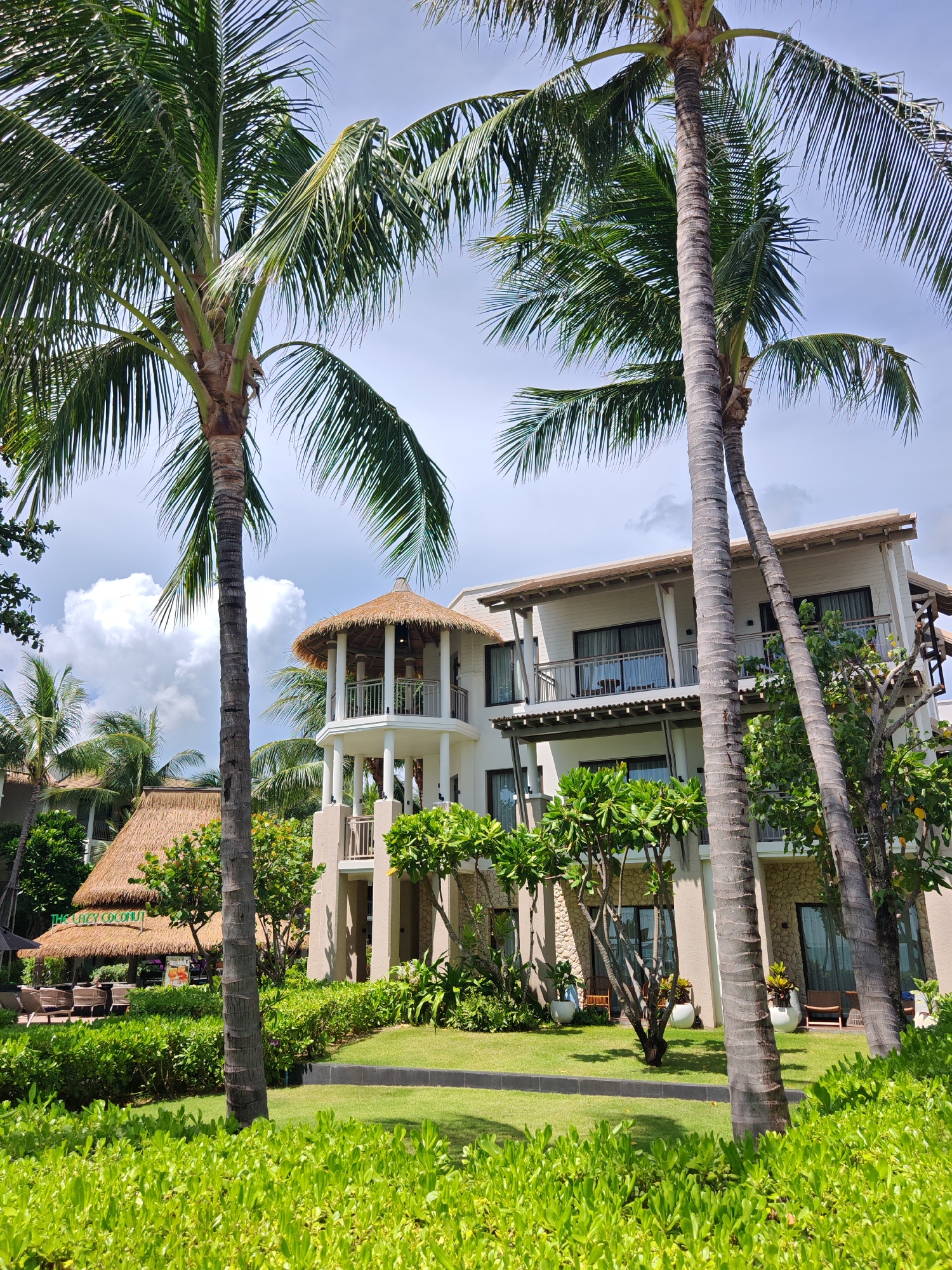 A resort complex, likely in Baan Bo Phut, Thailand, is depicted on a bright, sunny day with scattered clouds. The foreground is dominated by lush, green manicured bushes and a well-kept lawn. Two prominent palm trees, with rough, textured trunks and full fronds, frame the view.
Behind the palm trees, a modern, multi-story building with a light-colored facade and balconies is visible. The building has a contemporary architectural style with elements like stone accents and wooden trim. To the left of the main building, a smaller structure with a thatched roof, resembling a restaurant or bar, is partially obscured by greenery and another palm tree. This structure has outdoor seating areas, with tables and chairs visible. Another thatched-roof structure, possibly part of the resort's amenities, is situated further in the background. The overall impression is one of a tropical vacation destination, with a blend of natural elements and resort infrastructure. No people are visible in the image.