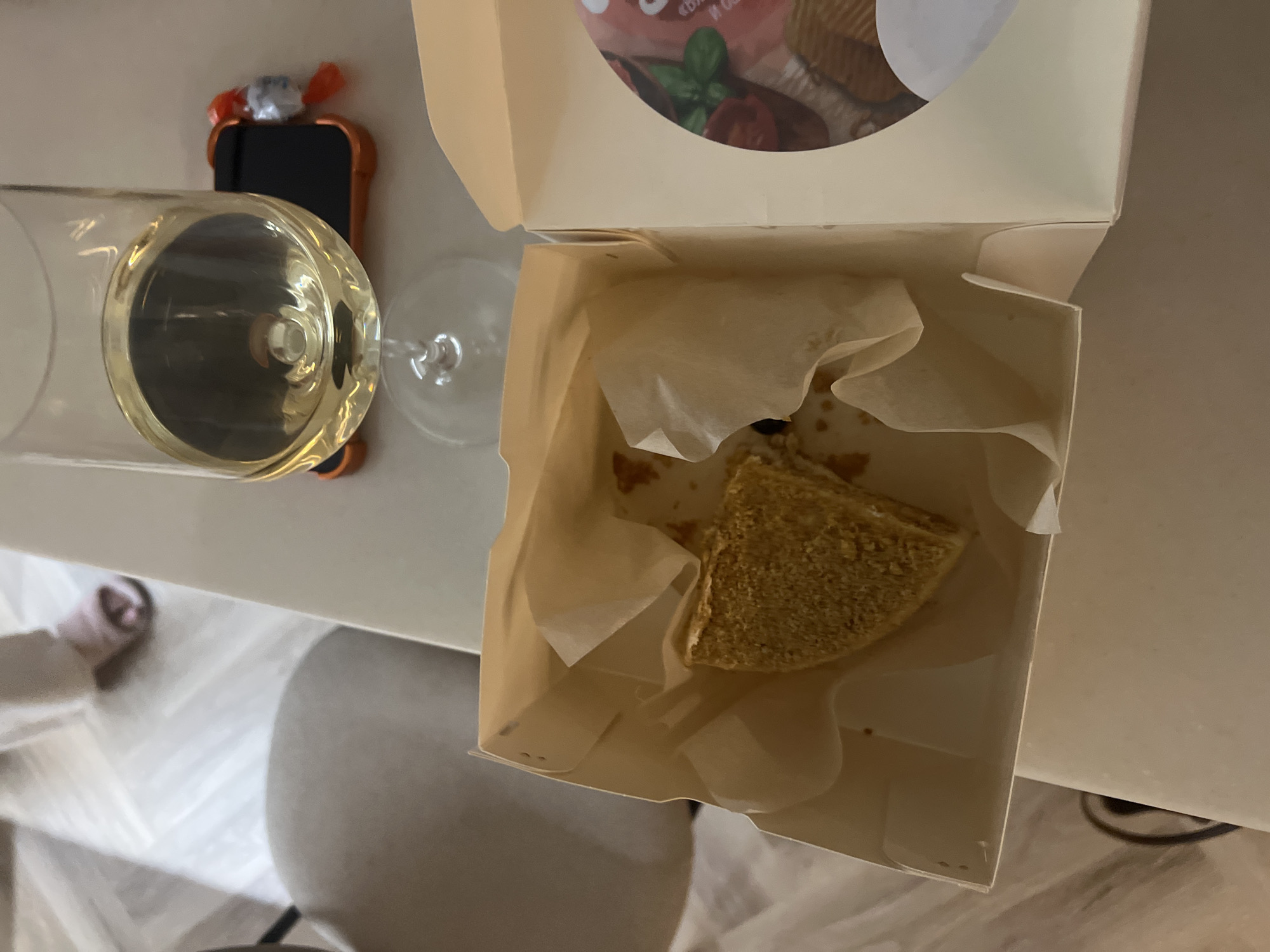 A high-angle shot shows an open take-out container with a partially eaten triangular piece of cake inside. The cake is light brown with a textured surface. The container is lined with parchment paper. To the left of the container is a partially filled wine glass with a clear, pale liquid. Behind the wine glass, there is a smartphone in a bright orange case. A white countertop or table is visible. A glimpse of a person's slippered foot can be seen on the bottom left. The setting is indoors, likely in a home, and the location is presumably in Рубцово, Russia, though this cannot be confirmed solely from the image.
