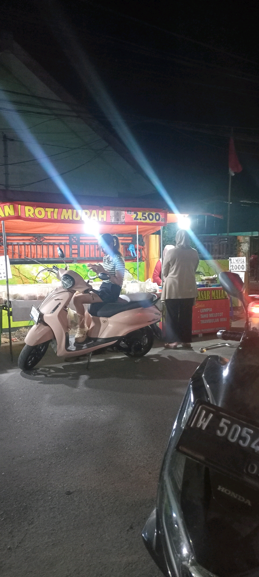 A horizontal shot shows a pink scooter parked on a paved road at night. A person wearing a striped shirt and dark shorts is seated on the scooter, looking at their phone. Behind the scooter is a food stall with an orange awning that reads "ROTI MURAH" in yellow letters. A price of "2.500 an" is visible. A woman wearing a headscarf and a long, light-colored tunic stands at the food stall, facing away from the camera. Another person in a red shirt is partially visible behind her. The stall has a colorful mural of trees and a green fence. To the right of the stall, another food stall sign reads "PASAR MALAM" with a list of food items including "LUMPIA", "TAHU MELOTOT", and "TRAMBULAN MINI". The foreground shows the front of another dark-colored scooter, with a license plate displaying "W 5054". The overall lighting is dim, with bright streaks of light from overhead sources. The location is identified as Bendotretek, Indonesia.