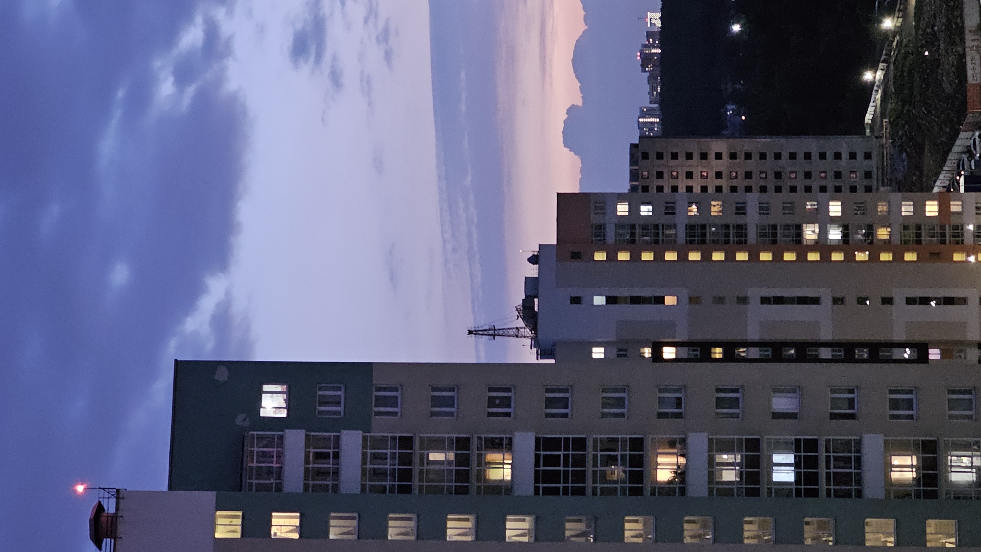 The image depicts a cluster of high-rise residential buildings against a twilight sky in Yekaterinburg, Russia. The sky is a gradient of deep blue and purple, with scattered clouds. The foreground is dominated by the facades of several apartment buildings, characterized by rows of windows, many of which are illuminated from within, suggesting occupancy. Balconies are visible on some of the buildings. One building on the left features a teal and pale yellow color scheme, while adjacent buildings display beige and orange tones.
On top of one of the buildings, a red aviation warning light is visible. In the middle ground, the silhouette of a construction crane is identifiable against the sky, indicating ongoing development or construction in the area. Further in the background, the distant city skyline is faintly illuminated by lights, and a dark, forested hillside is discernible. The overall scene conveys a sense of urban life at dusk. There are no people or specific actions explicitly depicted in the image. The environment is urban, with a mix of residential structures and infrastructure.