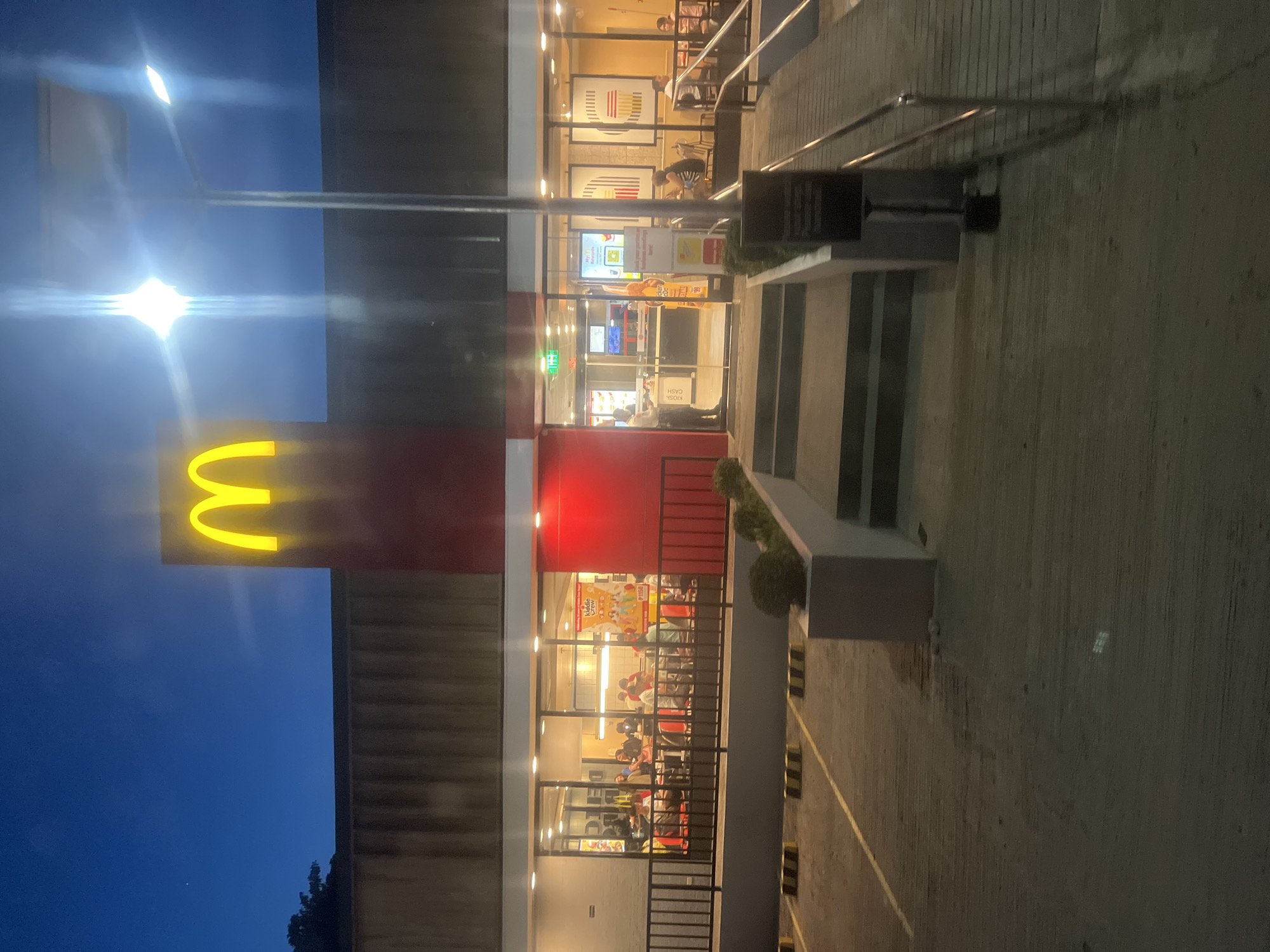 The image shows a McDonald's restaurant at dusk or night. The iconic golden arches logo is prominently displayed on a tall red and white pylon in front of the building. The restaurant has large glass windows, allowing a view of the interior where customers are seated at tables. The exterior features a parking lot with marked spaces and a ramp leading to the entrance, indicating accessibility. A street light illuminates the scene. The overall setting appears to be an urban or suburban area, and based on contextual clues like the currency (P150) on a poster, it is likely located in the Philippines, specifically Davao City, Philippines.