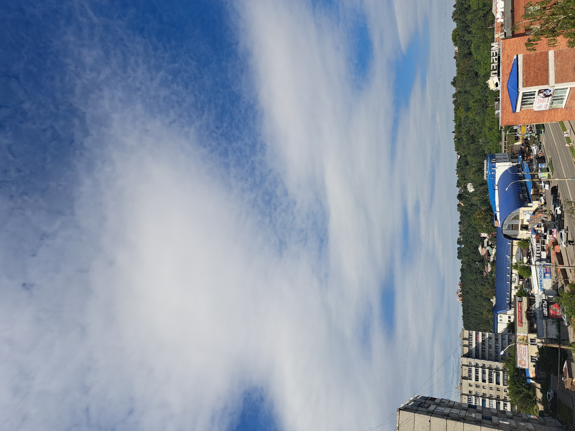 The image is a wide-angle, slightly tilted aerial view of a street scene in Irkutsk, Russia. The sky is a vibrant blue with scattered white, fluffy clouds. In the foreground, on the left, is a section of a grey, multi-story apartment building with visible balconies. A road runs horizontally through the middle ground, bordered by various commercial buildings. Some of these buildings have blue roofs, and signs in Cyrillic are visible, including "ЦВЕТЫ" (Flowers) and "ФАРМЭКОНОМ" (PharmEconomy). A few vehicles are present on the road, and several pedestrians are walking along the sidewalk. To the right of the road, another brick building with a blue-roofed extension is prominent. In the background, a dense, green forested hill rises, with scattered houses visible amongst the trees. The overall impression is of a typical urban landscape on a partly cloudy day.
