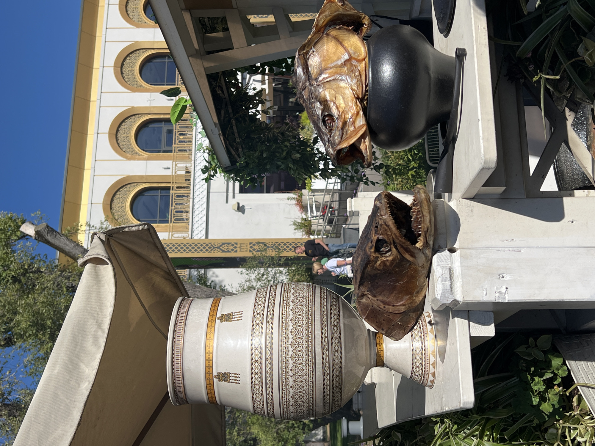 The image depicts a sunny outdoor scene in Moscow, Russia, with a prominent, large decorative vase featuring intricate patterns of gold and brown. In front of the vase, two preserved fish heads are displayed. One fish head, with its mouth open revealing sharp teeth, is positioned to the left, partially obscured by the vase. Another, larger fish head, with a golden-brown hue and visible gills, sits on top of a black, round pot to the right.
Behind these objects, a white structure with a canvas awning extends from the left side of the frame. Further back, a building with white walls and arched golden windows is visible, suggesting an establishment or cultural site. Two individuals, a man and a woman, are seen in the background, standing near a railing and appearing to converse. Lush green foliage surrounds the foreground elements, indicating a well-maintained outdoor space. The overall setting suggests a public area, possibly a market, garden, or exhibition space, with a blend of decorative elements and natural surroundings.