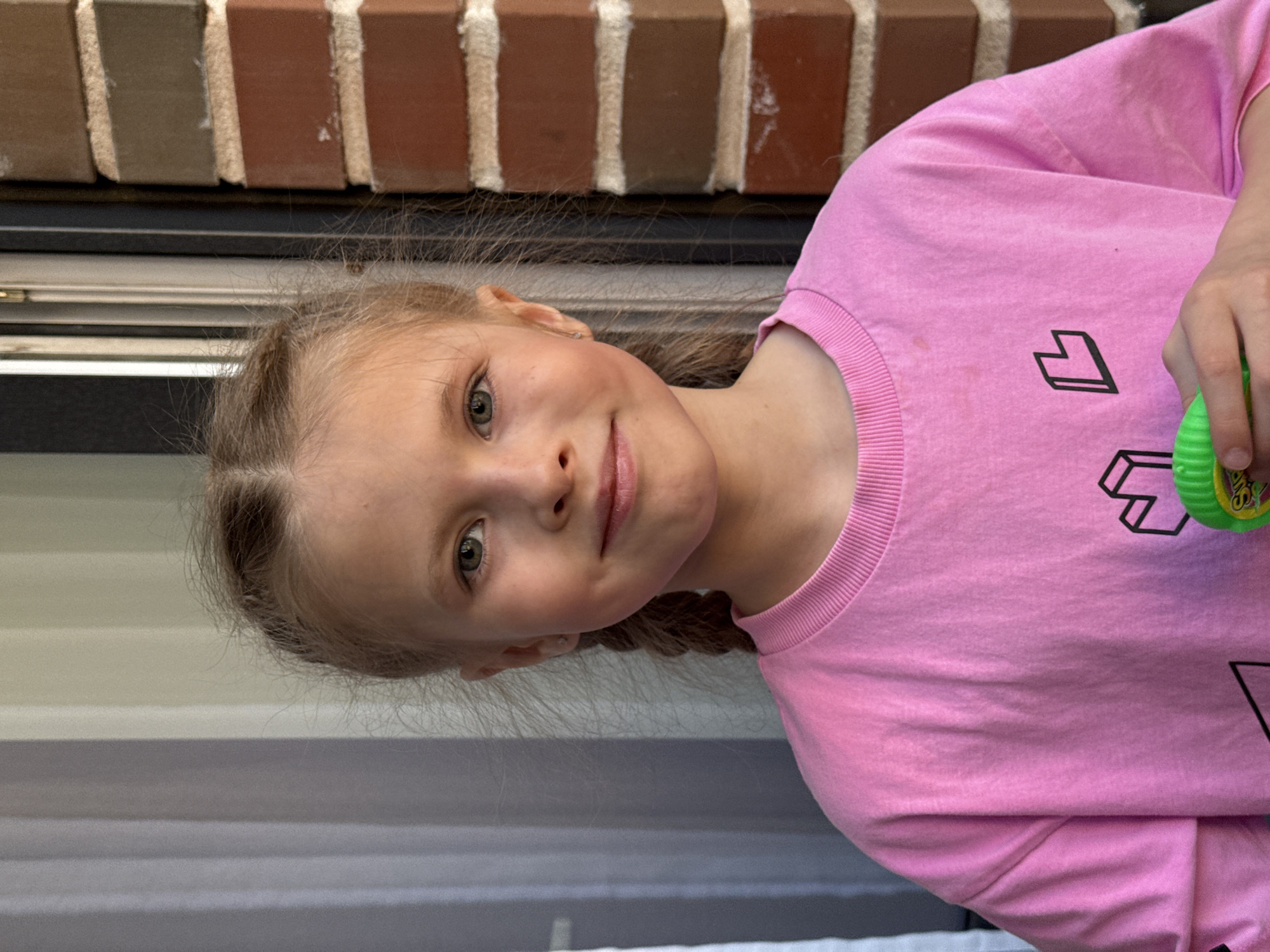 A young girl with blonde hair braided into pigtails is centered in the frame. She has fair skin and light eyes. She is wearing a pink long-sleeved shirt with black geometric designs on the front. The background consists of a brick wall with light-colored mortar and grey siding below. The location is not identifiable as Izhevsk, Russia. The girl is holding a green object in her right hand.