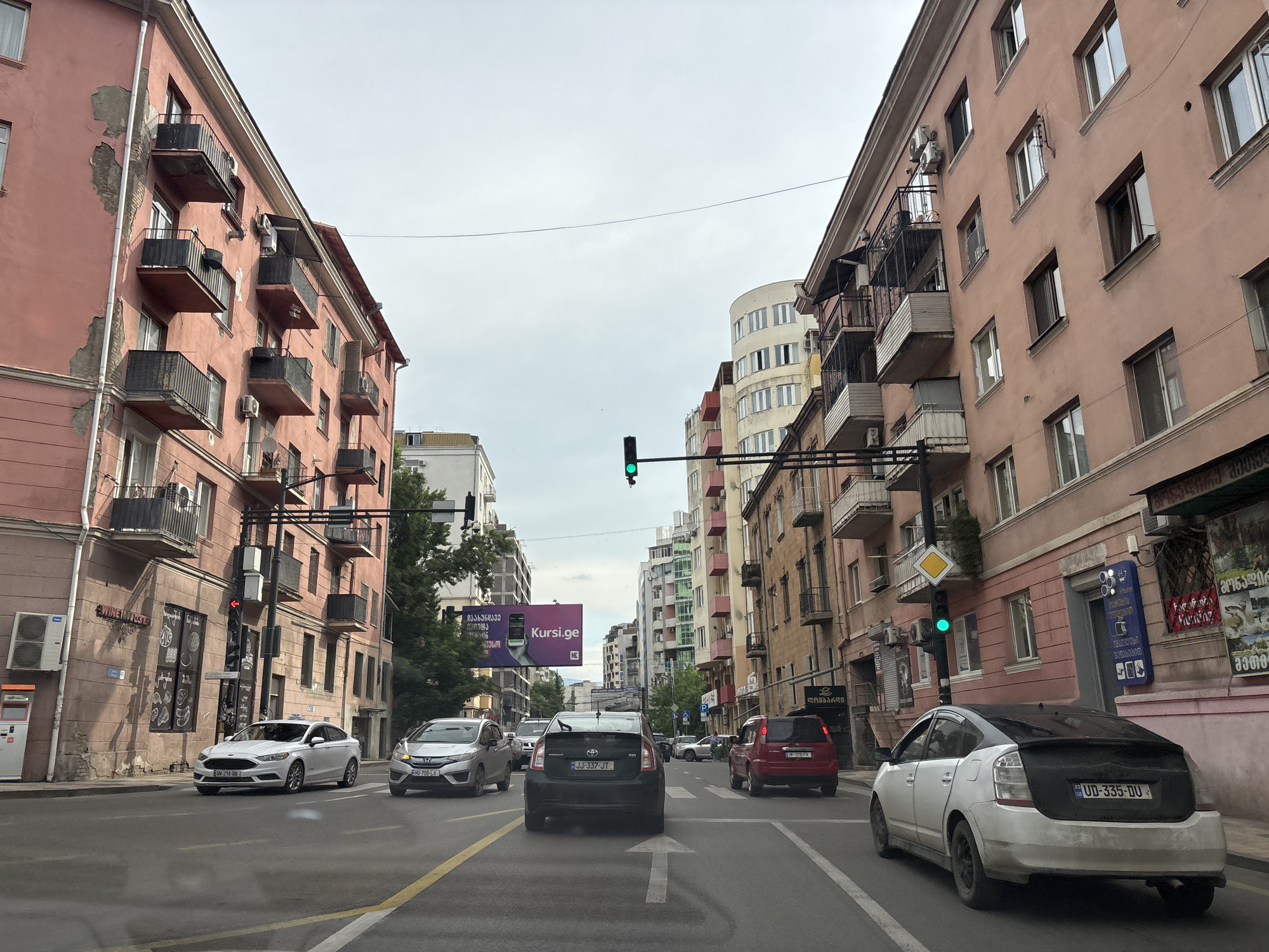 The image shows a street scene in Tbilisi, Georgia. The perspective is from a vehicle traveling on a multi-lane road. On either side of the street are multi-story buildings with balconies. The buildings on the left are painted a salmon pink, while the buildings on the right are a mixture of pink, peach, and yellow. There are several cars on the road, including a white Toyota Prius in the foreground on the right, and a black Toyota Prius behind it. A red SUV is visible further down the road on the right. A traffic light is visible, currently showing green. There are advertisements and signs on the buildings. The sky is overcast.
