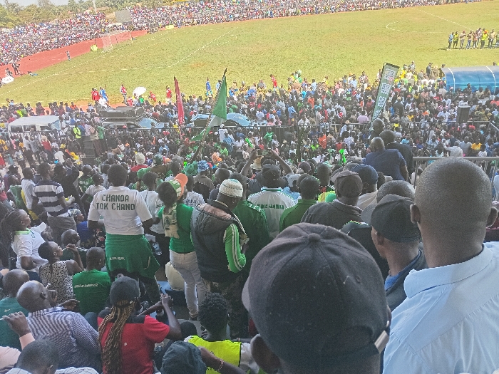 The image depicts a large, vibrant crowd gathered in an outdoor stadium under bright daylight, likely in Siaya, Kenya. The vast number of people fill both the stands and the field level, extending into the distance. Many attendees are dressed in green and white clothing, including shirts, scarves, and hats, and several green flags are prominently waved, suggesting a common affiliation or group identity. A red-colored running track encircles a green grass field, typical of a sports arena, with a blue dugout or shelter visible near the field's edge.
In the mid-ground, a person wearing a white t-shirt clearly displays the text "CHANDA TOK CHANO" on their back. Other visible objects include speaker systems or sound equipment mounted on a platform amidst the crowd, and a green banner with graphics, though its text is not legible. The weather appears sunny, contributing to the clear and bright illumination of the scene. The overall impression is one of a lively and well-attended public event, potentially a political rally, sporting event, or other large community gathering, characterized by a strong sense of collective enthusiasm and solidarity.