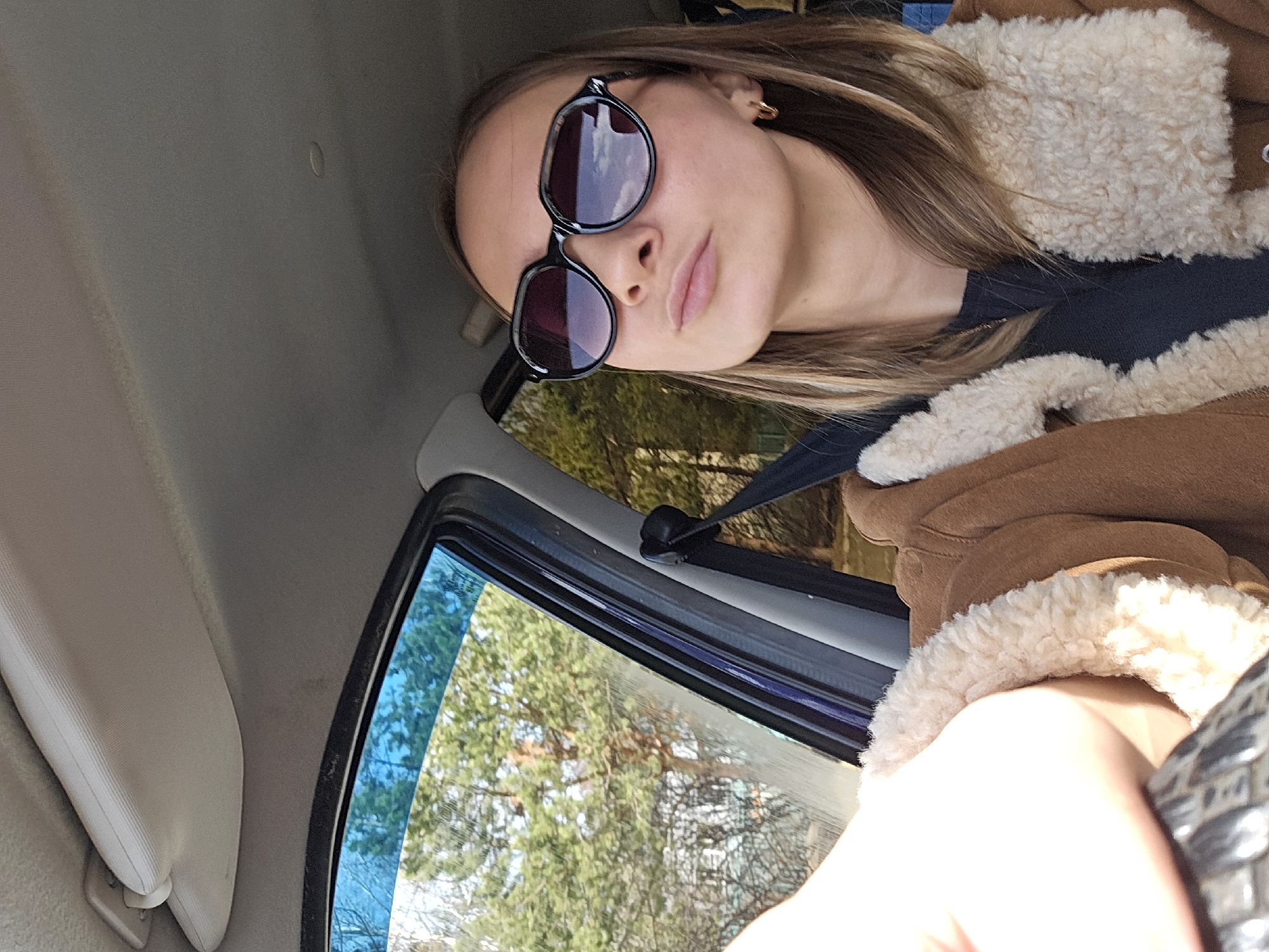 A woman is seated in a vehicle, likely the driver's seat. She is wearing sunglasses and a brown coat with a light-colored fluffy trim. She is wearing a seatbelt. The window shows a view of trees, suggesting a location with greenery. It is not possible to confirm the location as Chaikovsky, Russia, or provide further detail regarding the environment based on the single image.