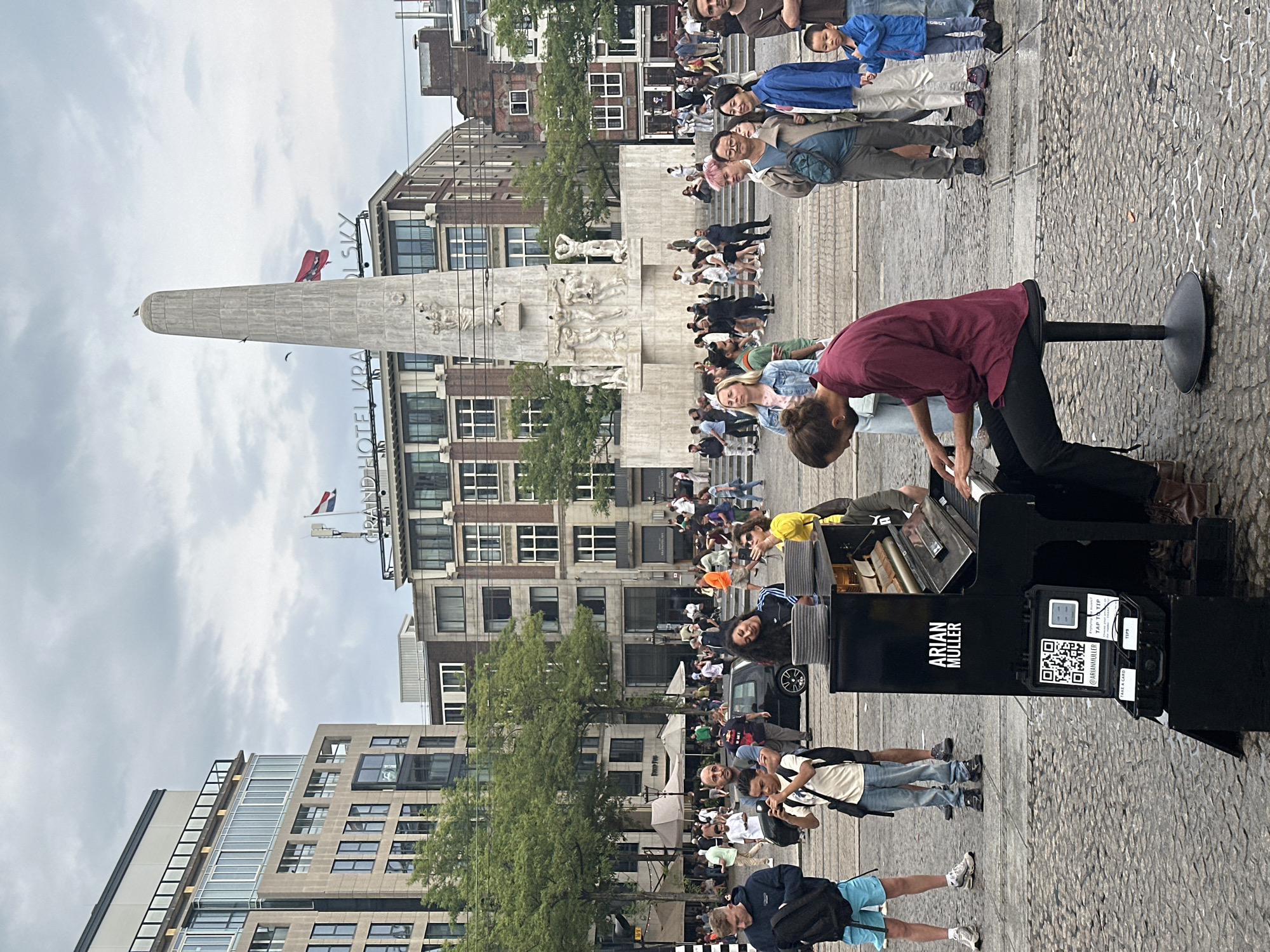 The image depicts Dam Square in Amsterdam, Netherlands, under a cloudy sky. A man is playing a portable piano on the cobblestone ground, with the National Monument and the Grand Hotel Krasnapolsky visible in the background. The pianist is seated on a black stool, wearing a burgundy shirt and black pants. The piano has "ARIAN MULLER" written on it, and a QR code and payment information are displayed for tips. A crowd of people, some watching the performance and others walking by, surrounds the pianist. Among the crowd, a young boy in a blue jacket stands near the piano, looking on. The scene captures a moment of public performance and urban life in Amsterdam.