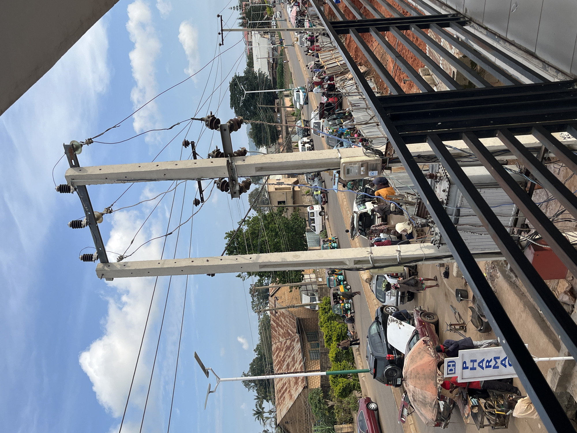 The image captures a busy street scene in Bukuru, Nigeria, with a focus on a section of road where vehicles and pedestrians are interacting. Prominent power lines and concrete utility poles dominate the upper portion of the frame, indicating the presence of essential infrastructure. In the foreground, a black metal railing overlooks the street, suggesting the vantage point is from an elevated structure or walkway.
On the street below, a variety of vehicles are present, including cars, a black SUV with its hood open, and several auto-rickshaws, commonly known as Keke Napep. Some of these vehicles appear to be parked or stopped, with people gathered around them. One car, a maroon sedan, is parked with its trunk open. A makeshift stall with an umbrella is set up next to another car, with individuals attending to it. A blue and white sign with the text "DH PHARMA" is visible near this stall.
Pedestrians are seen walking along the road and sidewalk, some carrying items, suggesting daily commerce or movement. The environment appears to be a built-up urban area with buildings lining the street, characterized by corrugated iron roofs on some structures and brickwork on others. Green foliage and trees are interspersed throughout the scene, indicating a degree of greenery within the city. The sky is partly cloudy with blue skies visible, suggesting daytime. The ground is paved or dirt in some areas, with a section of exposed red earth and construction materials behind the railing in the foreground. The overall impression is of a vibrant and active urban environment with various activities taking place on the street.