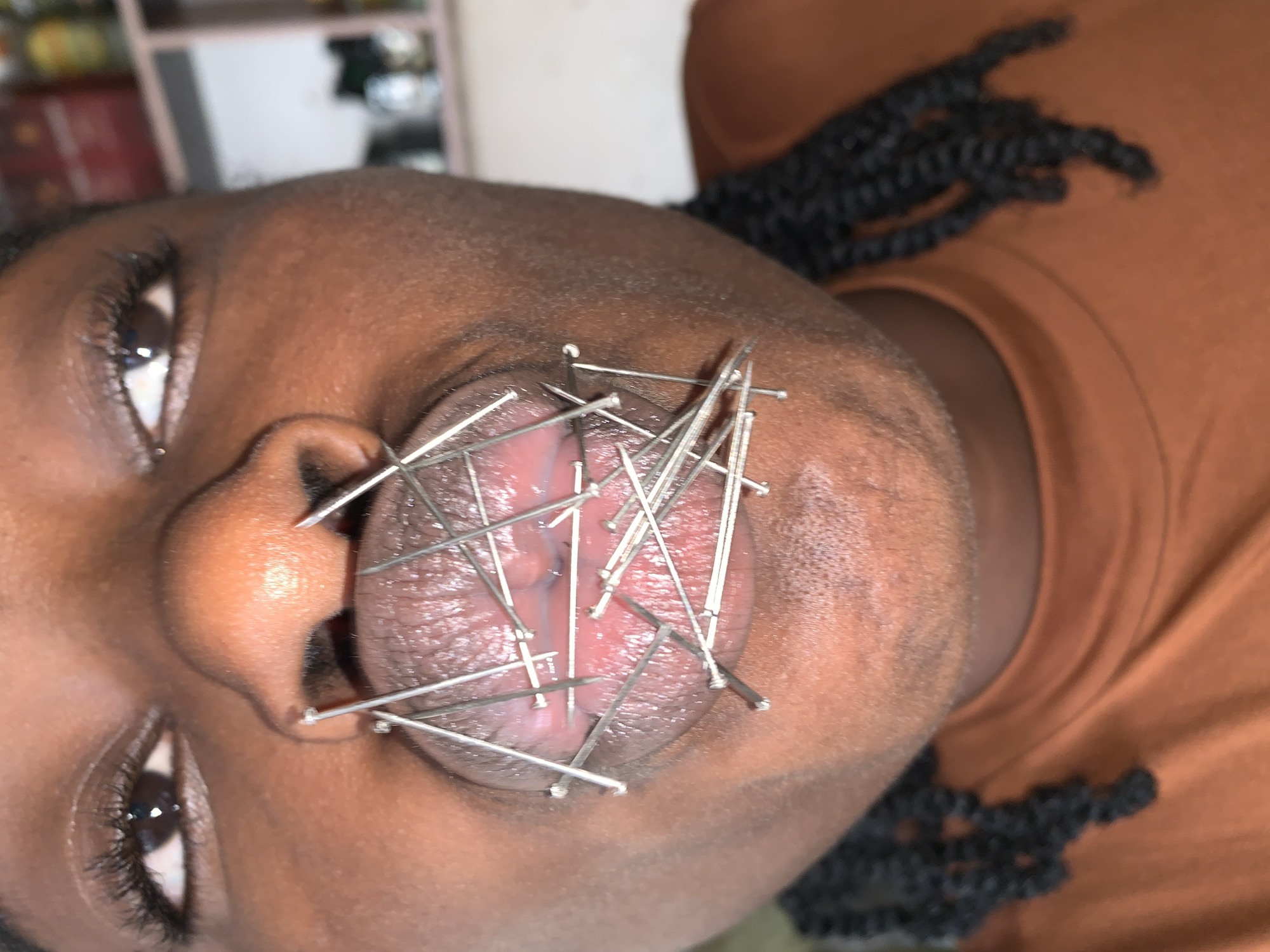 A close-up, eye-level forensic image taken in New Stadium, Nigeria, depicts an individual with dark skin, visible from the eyes to the upper chest. The individual has open eyes, dark eyelashes, and braided hair visible over the shoulders. They are wearing a terracotta-colored t-shirt. The central focus is the individual's wide-open mouth, revealing the tongue. Numerous metallic, needle-like pins, each with a flattened head, are inserted into and through the tongue and surrounding oral tissues, forming an irregular, crisscrossing pattern. The pins appear to hold the mouth open and the tongue perforated. Reflections are present on the metallic surfaces of the pins, indicating an external light source. The background is out of focus, showing indistinct shelving with various items on the right side.