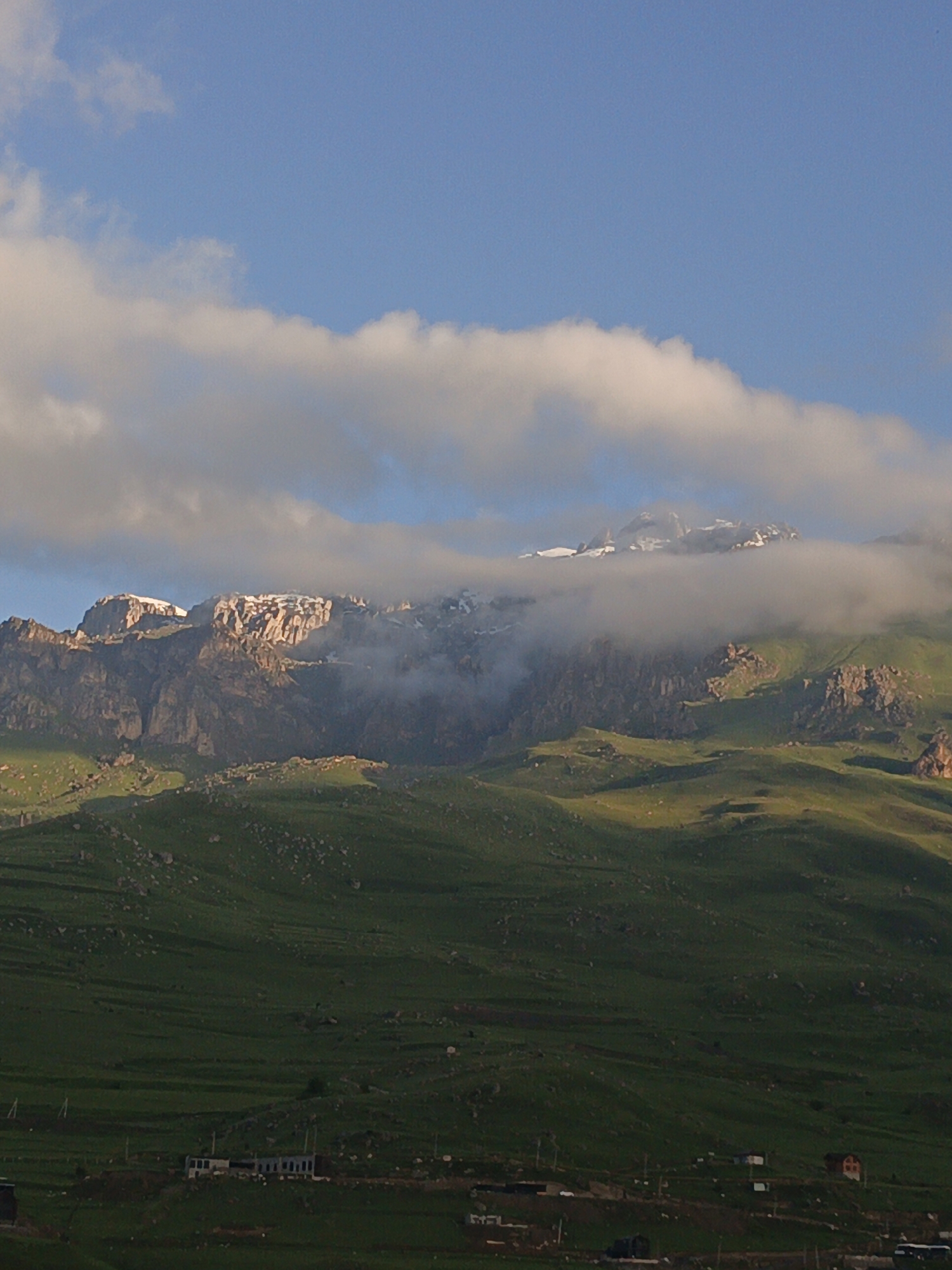 The image depicts a wide, panoramic view of a mountainous landscape near Verkhny Fiagdon, Russia. The foreground features rolling green hills dotted with scattered rocks, transitioning into steeper, rocky mountain slopes. A thin layer of clouds partially obscures the peaks of the majestic mountains in the background, some of which are capped with snow. The sky above is a clear blue, with some scattered white clouds. At the base of the hills, several small buildings are visible, suggesting a settlement. The overall scene is one of natural beauty and serene wilderness.