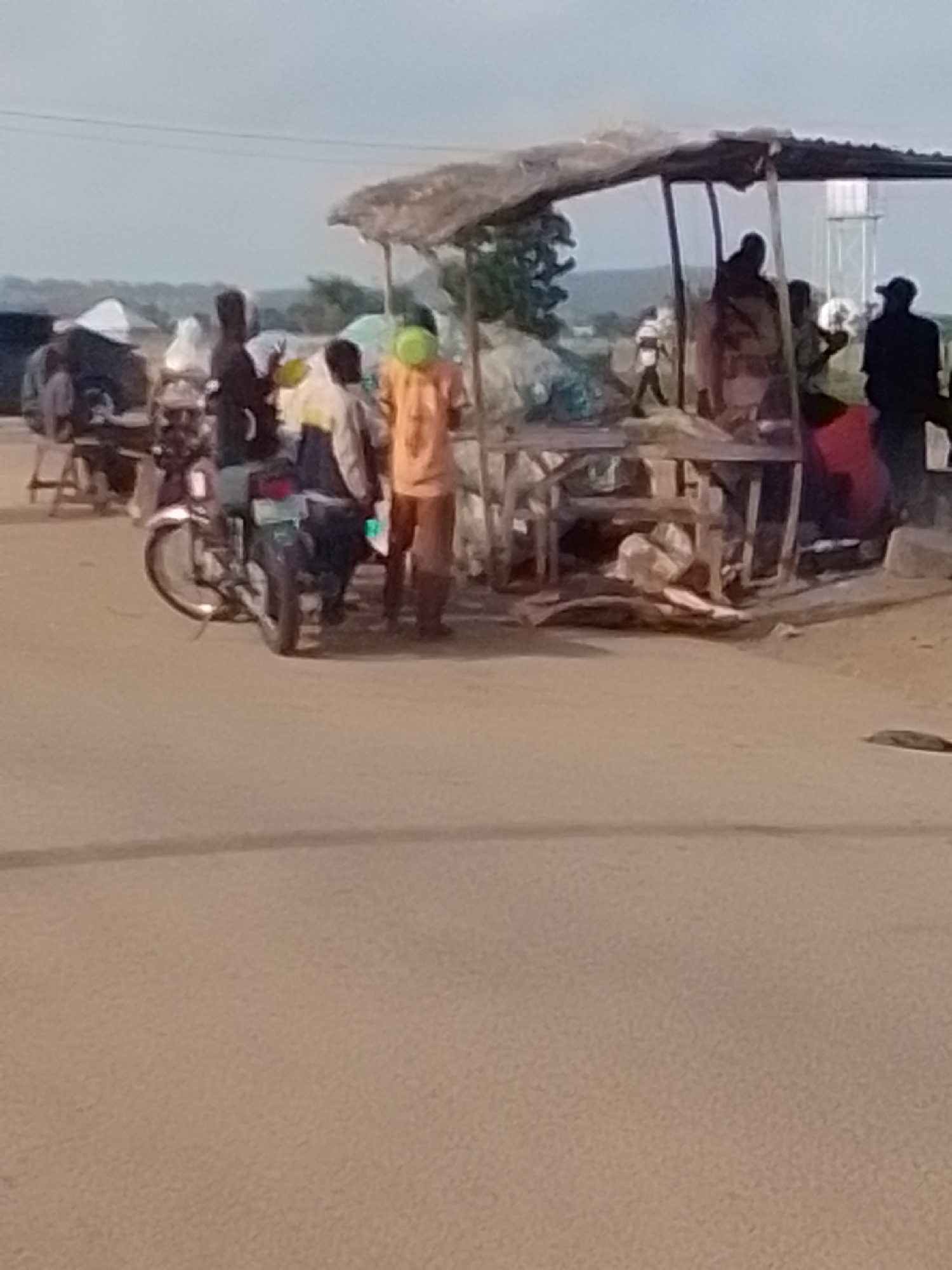 This image depicts an outdoor market scene in Gombe, Nigeria. A group of people are gathered around a makeshift stall covered by a thatched roof. The stall appears to be selling goods, with various items displayed on wooden tables and on the ground. A motorcycle is parked to the left of the stall, with a person seated on it. Several individuals are standing or walking around the stall, some interacting with each other. The background shows a hazy sky and a distant landscape, possibly indicating a semi-urban or rural setting on the outskirts of Gombe city. The ground is paved with a rough surface, likely a road or dirt path common in the region. The overall atmosphere suggests a busy day at the local market.