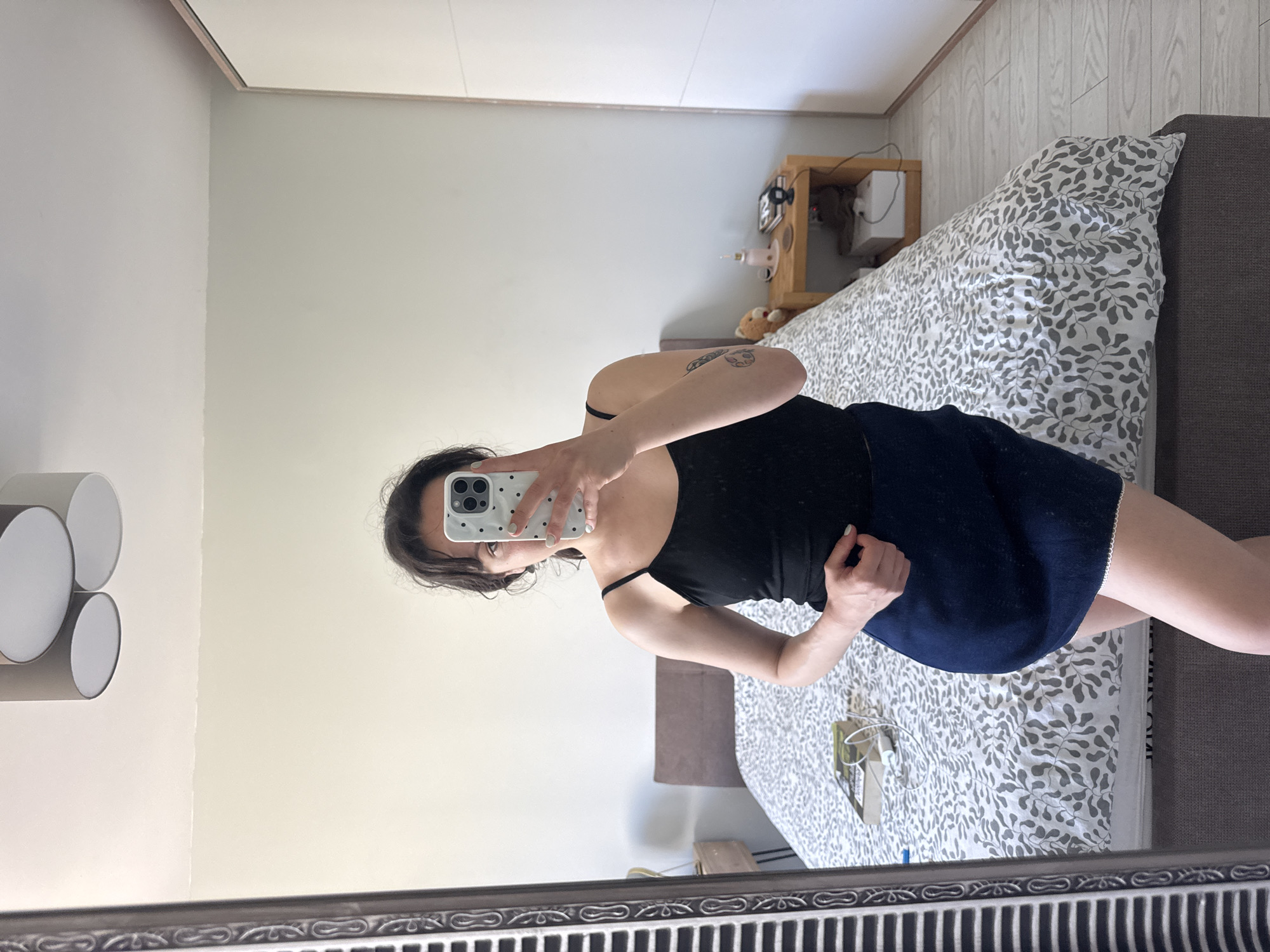 A mirror selfie captures a woman standing in what appears to be a bedroom in Burgas, Bulgaria. The woman is reflected from the chest down, wearing a black spaghetti-strap top and a dark blue skirt with a white lace trim. Her left arm is raised, holding a white smartphone with a white and black polka-dot case, obscuring her face. Her right hand rests on her hip. A circular tattoo is visible on her upper left arm. She has dark hair and light nail polish.
The background shows a neatly made bed with a gray headboard and a duvet cover featuring a white background with a gray leaf-like pattern. To the right of the bed, a light brown wooden nightstand holds a small teddy bear, a light-colored lamp or diffuser, and some wires. The walls are light cream or off-white. On the wall to the left, a modern, multi-circular white light fixture is visible. The floor or lower wall area behind the bed features light-colored wooden paneling. The mirror itself has a decorative, dark-colored frame at the bottom. The lighting in the room appears soft and ambient, suggesting indoor lighting. No discernible text is visible in the image.