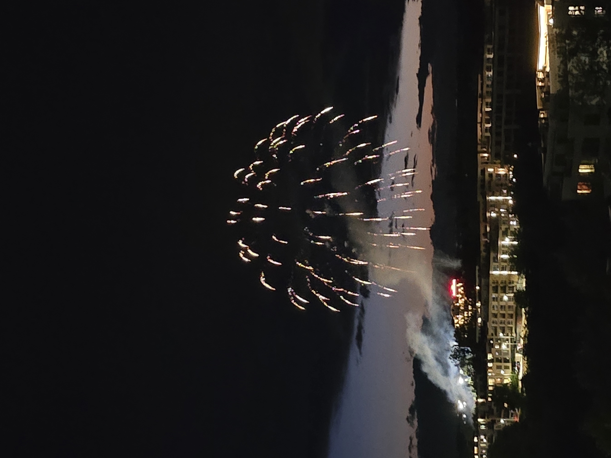 The image shows a nighttime scene with fireworks exploding over water. In the background, there are illuminated buildings, possibly a cityscape. The sky is dark, contrasting with the bright bursts of the fireworks. Smoke or haze is visible near the water and buildings. The scene appears to be a celebration or event taking place near a waterfront area with urban development.