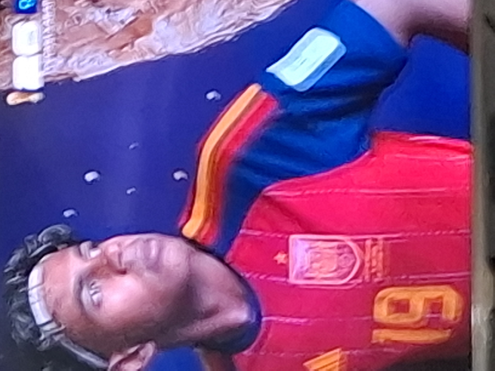 The image shows a close-up of a person wearing a red and blue jersey with the number "19" on it. The person appears to be a young male with curly hair and a white headband. The jersey has a crest on the chest that includes a star, and the sleeves have colorful stripes. The background is a dark blue with some white, blurry shapes. There is no discernible location in Almaty, Kazakhstan visible in this image.