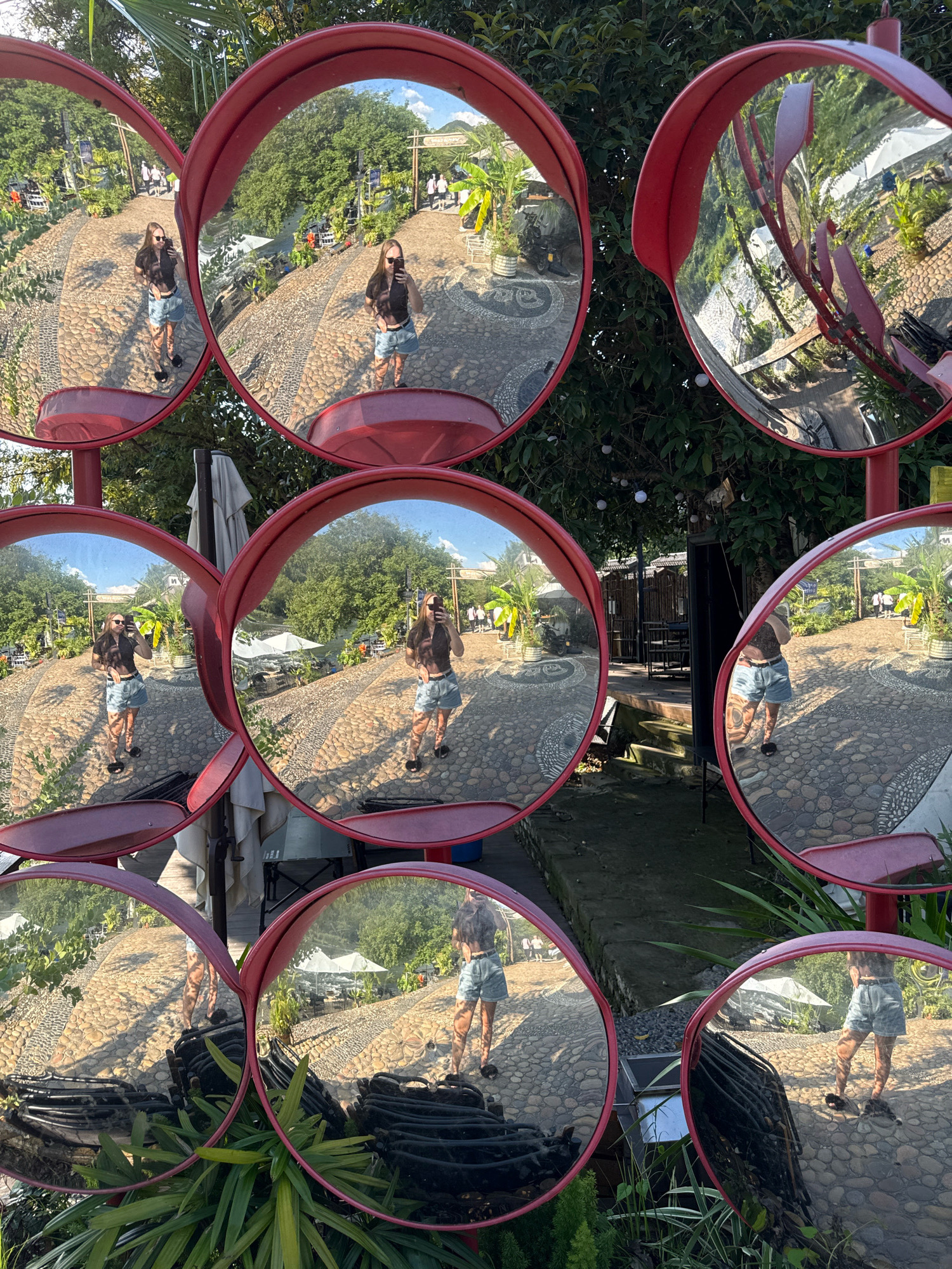 The image is a photographic composition featuring a series of convex security mirrors, likely located in Yiwu, China, given the context. Each mirror reflects distorted views of a paved outdoor area with a pebble-like surface and scattered cobblestones. Lush green foliage and trees are visible, along with some architectural elements such as a wooden structure and white umbrellas. In several of the mirror reflections, a young woman is captured taking a selfie. She is dressed in a dark top and denim shorts, and she is holding a smartphone up to take the photograph. The mirrors create multiple fragmented reflections of the woman and the surrounding environment, giving the image a complex and multi-perspectival quality. Some reflections show other people walking in the background, and hints of parked vehicles and outdoor seating arrangements are also discernible. The overall scene suggests a public outdoor space, possibly a park, garden, or commercial area, common in Yiwu.