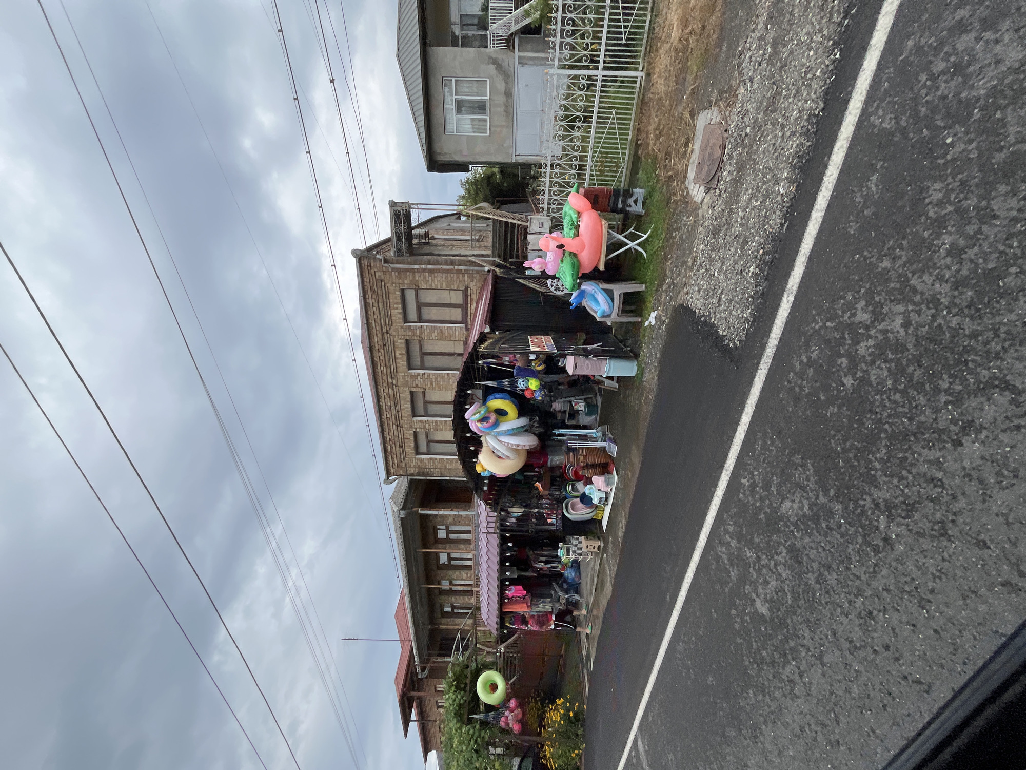 The image shows a street-side stall selling inflatable pool toys and other merchandise in Poti, Georgia. In the foreground, a wet asphalt road with a white line runs horizontally. The stall itself is set up in front of a two-story building with a brick facade and a tiled roof. The stall features a colorful assortment of items, including inflatable flamingos, unicorns, and rings, as well as beach balls and other beach accessories. A person, wearing shorts and a t-shirt, is seen browsing the merchandise. To the right of the stall, a white metal fence encloses a grassy area with a small building. The sky is overcast with clouds, suggesting a potential for rain. The scene depicts a typical roadside market in a coastal town.