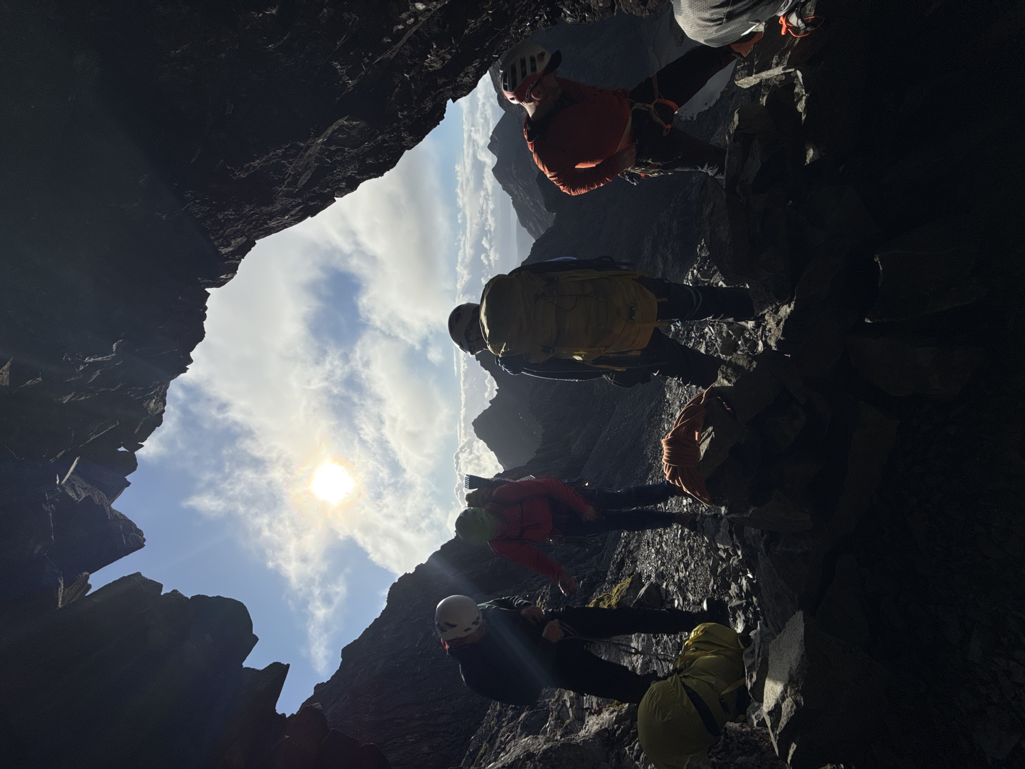 The image shows a group of people standing on a rocky terrain within what appears to be a cave or rock formation, likely in a mountainous area. The view is from inside the cave looking outwards, with the sky and a bright sun visible. The people are wearing outdoor clothing, including backpacks and helmets, suggesting they are engaged in some form of outdoor activity such as hiking or climbing. The foreground is dominated by the dark, jagged rock formations of the cave. The sky is visible in the distance. There is no location in the city specified.