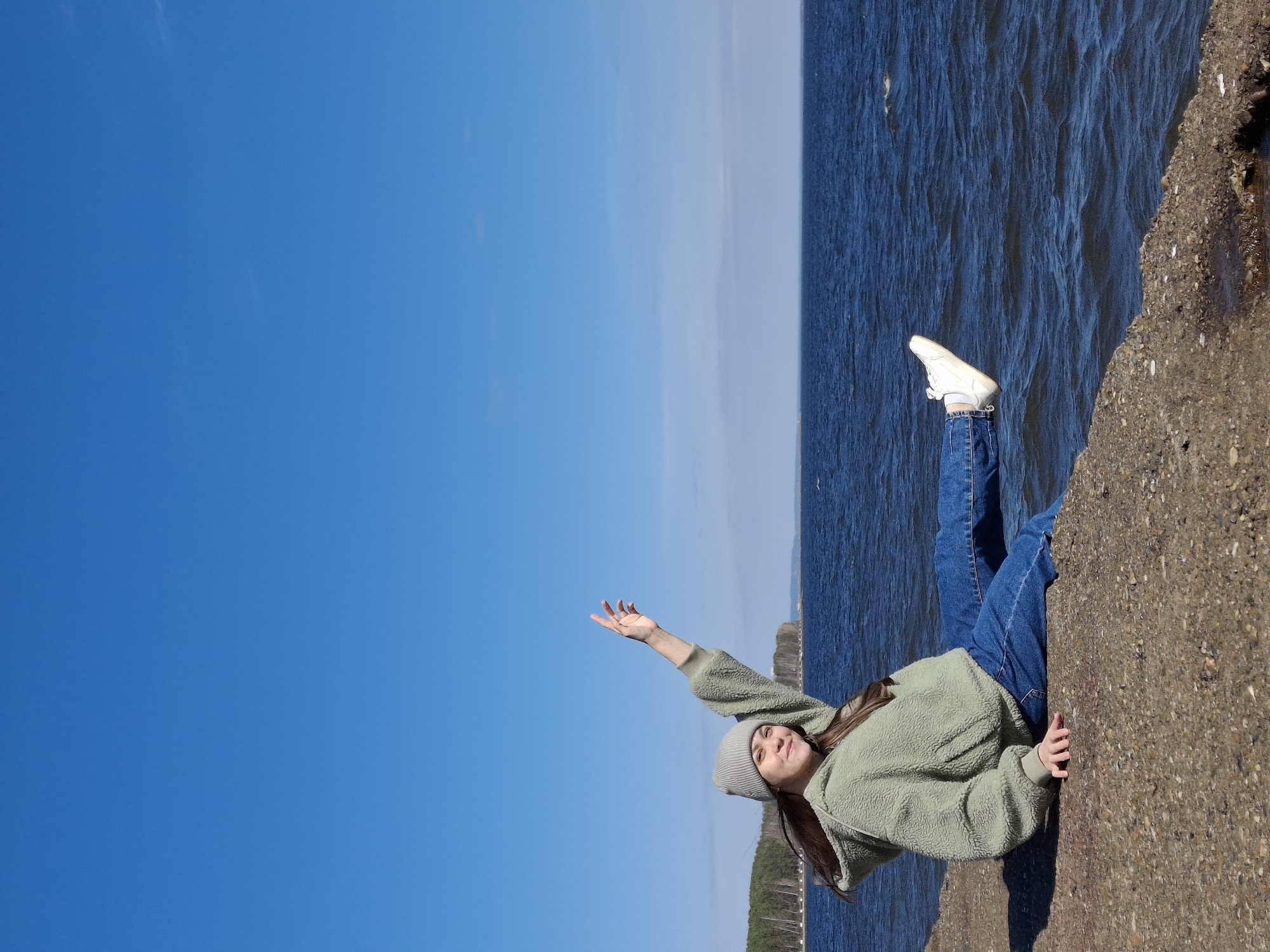 A young woman stands on a concrete structure adjacent to a body of water. She is wearing a gray beanie, a light green jacket, and blue jeans. Her right arm is raised towards the sky. The background features a clear blue sky above the water, which extends to the horizon. The scene is located in Chaikovsky, Russia.