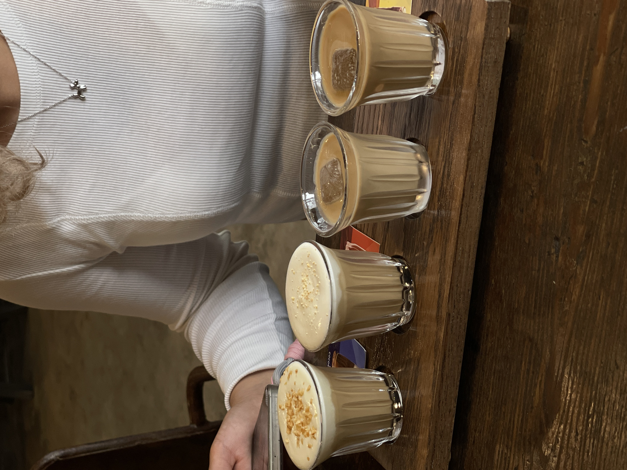 A close-up, rotated view depicts a person with fair skin wearing a white, ribbed long-sleeved shirt, sitting at a wooden table. They wear a silver necklace with a geometric pendant. A wooden tray holds four small glasses of a beige-colored beverage. The top two glasses contain ice cubes. The bottom two glasses are topped with a creamy white foam and garnished with what appear to be nuts. The location is possibly Da Lat, Vietnam, as it is known for coffee and the beverages resemble Vietnamese egg coffee or similar variations. The person appears to be holding a mobile phone.