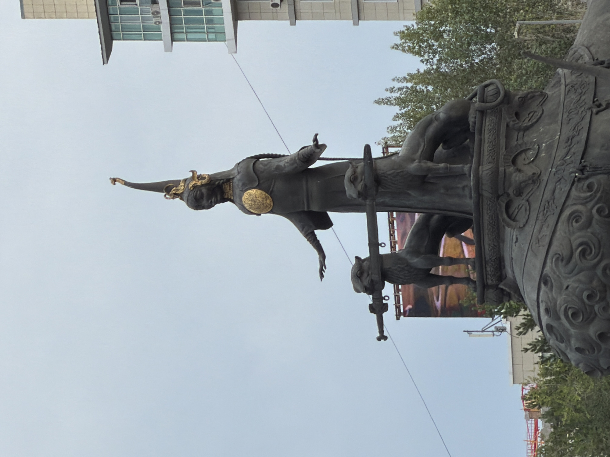 The image shows a bronze statue of a woman in traditional attire, wearing a tall, ornate hat with gold accents and a circular emblem on her chest. She is standing with her arms outstretched. Two bronze lion-like creatures flank her, each holding a long object horizontally in its mouth, resembling a sword or staff. The statue is mounted on a large, decorative base, which itself appears to be part of a larger monument. In the background, there is a modern building with a glass facade and a cloudy sky. To the left, a large billboard with an image of a winding road is visible. Trees are present in the mid-ground and foreground. This statue is identified as the "Mother of the Steppes" monument and is located in Astana, Kazakhstan.
