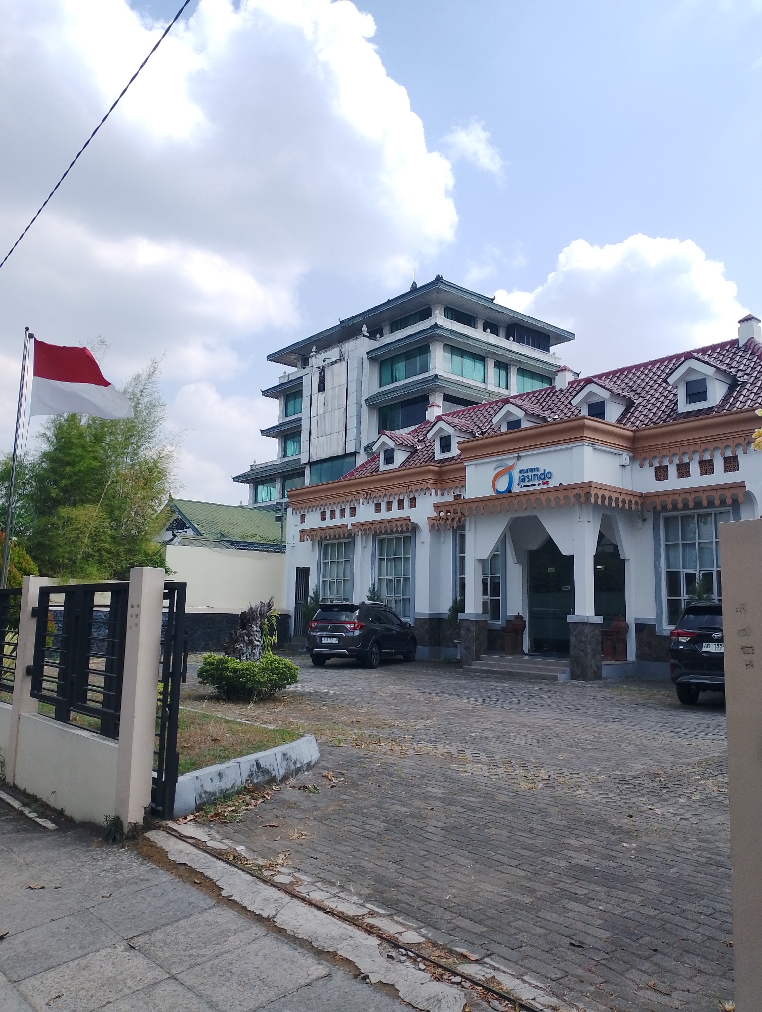 The image shows the exterior of the Jasindo Insurance office in Yogyakarta, Indonesia, on a partly cloudy day. The building features a modern multi-story structure behind a lower, older-style building with a red tiled roof and arched entrance. An Indonesian flag flies proudly on a pole to the left of the frame. The foreground consists of a paved driveway and sidewalk, with some scattered leaves. Two black cars are parked in front of the older building. Lush green trees and bamboo are visible behind the flag and fence. The overall impression is of a well-maintained office building in a tropical urban environment.