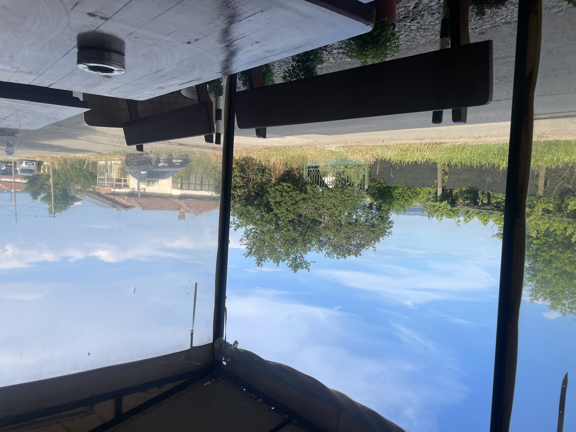 The image displays a daytime view from an outdoor seating area, likely a cafe or restaurant, located in Konstantinovo, Bulgaria. In the foreground, dark wooden tables and benches are visible. A silver-toned metal ashtray rests on the nearest table, reflecting ambient light. Small potted plants are situated on the ground beneath the benches. The view extends through a transparent plastic curtain or screen that serves as an outer wall for the seating area.
Outside, a paved road runs horizontally across the midground. To the left of the road, a grassy verge is present, bordered by a black mesh fence and further left by a green metal gate. Abundant green foliage, including trees and bushes, lines the road and fills the landscape. In the background, several houses with reddish-brown tiled roofs are discernible. A dark-colored SUV is parked or positioned near one house on the left side of the road, while two lighter-colored vehicles are visible further down the street on the right. The sky overhead is clear blue with sparse, elongated white clouds. No individuals are present in the scene.