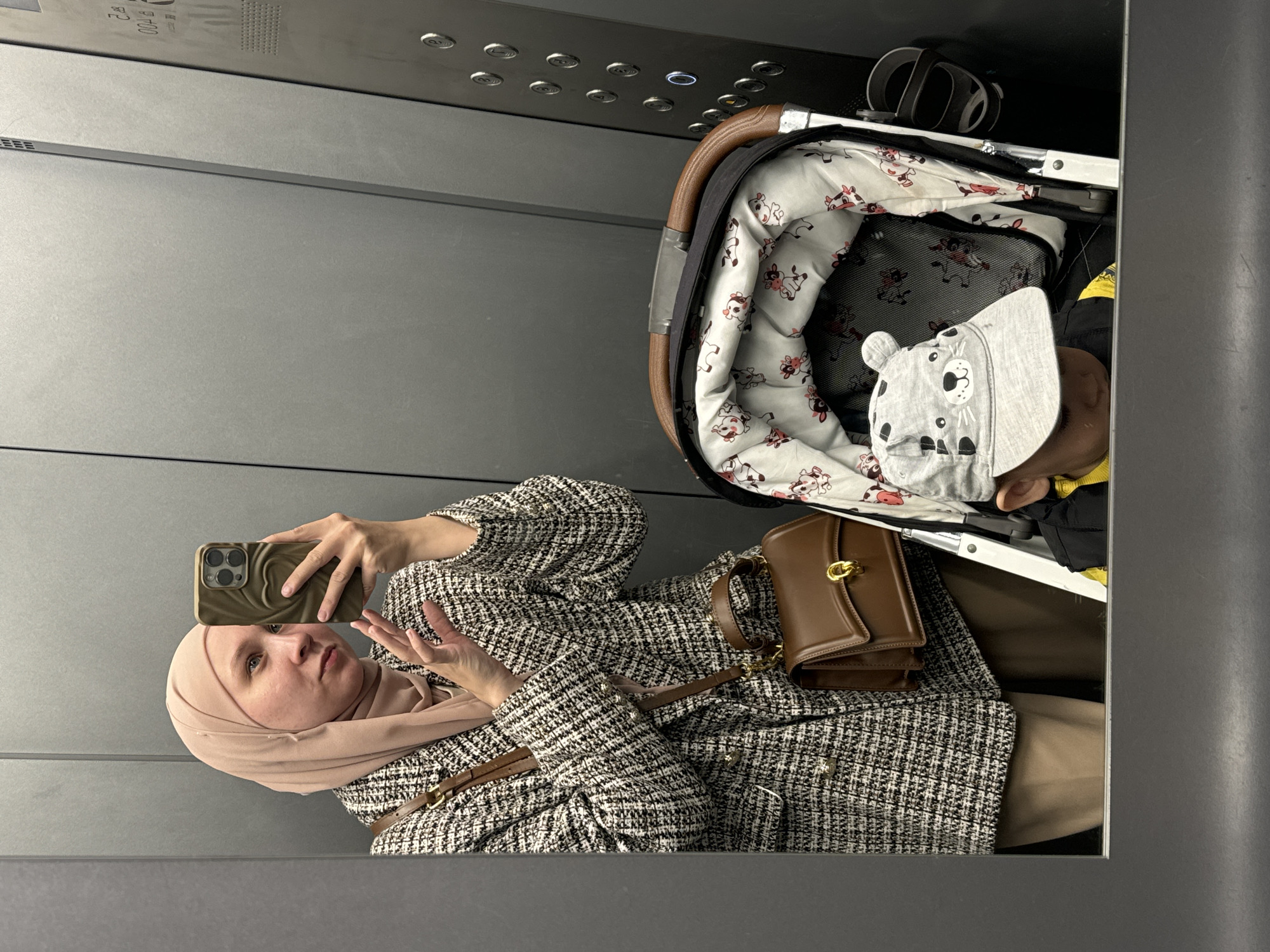 A mirror-viewed photograph captures a woman in a hijab taking a selfie in an elevator, with a baby in a stroller. The woman wears a tweed-patterned jacket, holds a smartphone in her left hand, and has a brown leather handbag over her jacket. A stroller with a baby lies to her right, the baby is wearing a patterned cap. The elevator's control panel is visible above the stroller. The scene is brightly lit. There is no verifiable information to indicate this photo was taken in Chaikovsky, Russia.