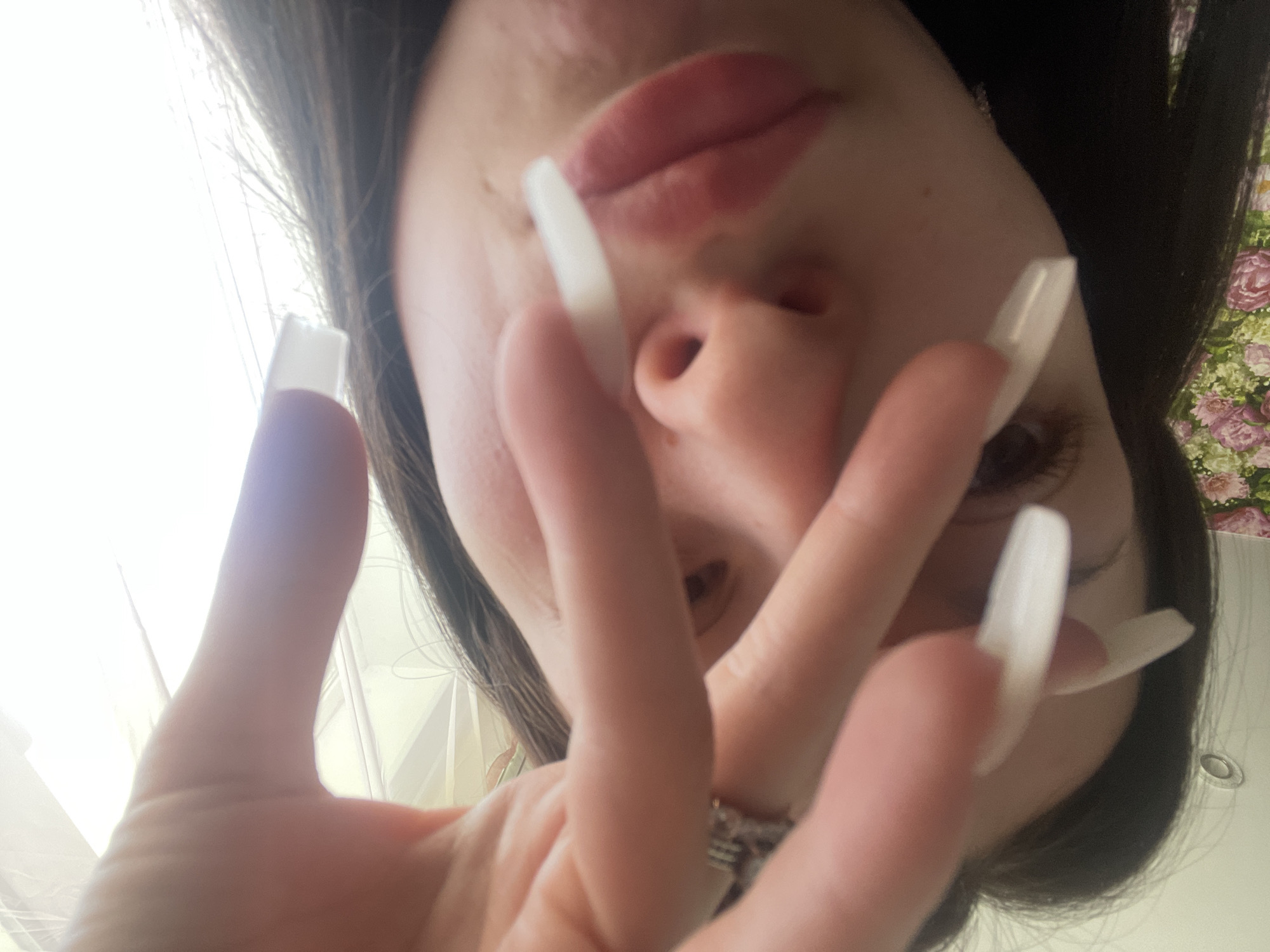 An upward-facing, close-up shot captures a person, likely female, from a low angle. The subject's face is partially visible at the top and center of the frame, with her hands and fingers prominent in the foreground, appearing large due to the close proximity and camera angle.
Her lips, which appear to have lip gloss or lipstick, are visible, along with parts of her nose and eyes. Dark hair frames her face. The most striking detail is the set of long, square-tipped fingernails, which are light-colored, possibly white or clear, extending prominently from her fingers. A ring can be seen on one of her fingers in the foreground.
The background is brightly lit on the left, suggesting a window or strong light source, indicating it is likely daytime. On the right side of the frame, a floral-patterned fabric, possibly part of bedding or decor, is visible. One of the cropped images also reveals a faint, circular, metallic object in the bottom right, which could be a drain or a light fixture, hinting at an indoor setting, possibly a bathroom or bedroom.
The person appears to be posing, perhaps intentionally showcasing her nails. No discernible text is visible in the image.