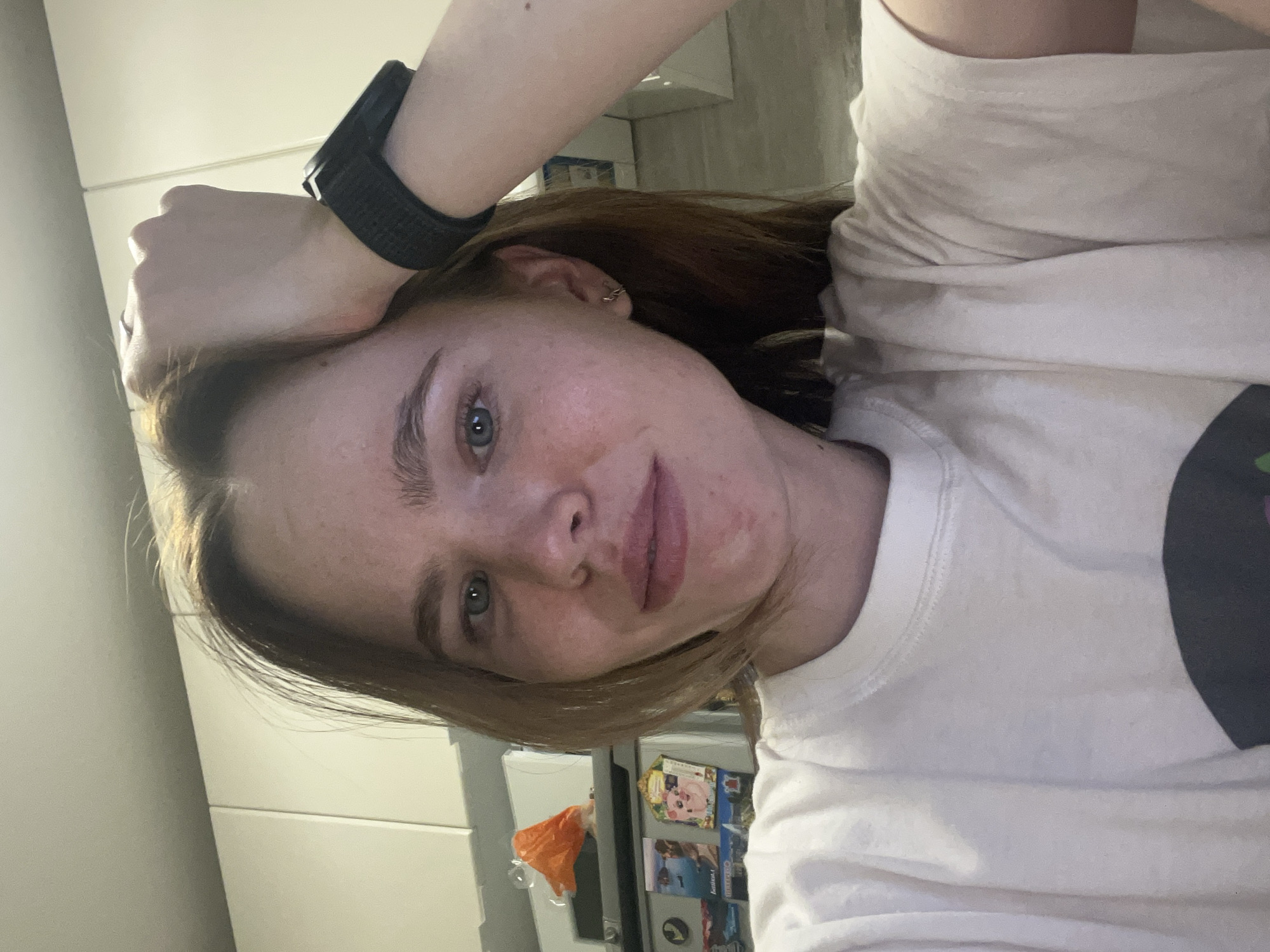 A selfie shows a young woman with light skin, light brown hair, and blue eyes. She wears a black fitness tracker on her left wrist and a white t-shirt. Her head rests on her left hand. In the background is a white refrigerator with magnets and what appears to be a white cabinet or wall. The location is likely an indoor kitchen space, but the specific location in Chaikovsky, Russia, cannot be determined from the image alone.