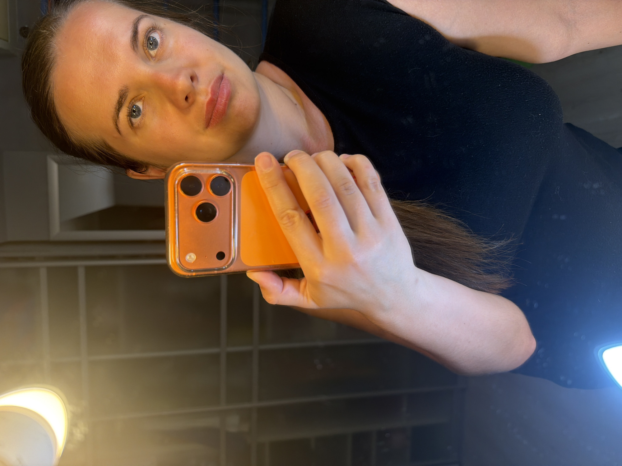 A woman takes a mirror selfie indoors in Sochi, Russia, captured in a medium shot. She has fair skin, light blue-grey eyes, and her long dark brown hair is pulled back, revealing her face. She looks directly into the camera with a calm, neutral expression, her lips tinted with a natural-toned lipstick.
She holds an orange or rose gold smartphone, likely an iPhone due to its visible triple-lens camera array, in her right hand. Her hand and arm are visible, with her fingers wrapped around the phone's casing. She is wearing a black top, possibly a t-shirt, which appears to be made of a slightly textured fabric.
The reflection is cast upon a dark, glossy surface, presumably a mirror or highly reflective wall, which shows faint linear patterns in the background. Two bright, artificial light sources are prominently reflected in the lower left and right areas of the image, illuminating the scene. No discernible text is visible.