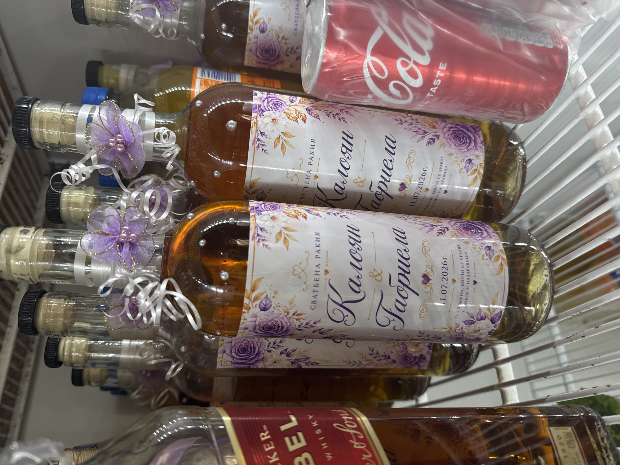 The image shows a close-up view of several bottles and a can of Coca-Cola on a white metal shelf, likely in a refrigerator or pantry. The bottles on the right are clear glass, filled with a golden liquid, and are decorated with purple and white floral labels. The labels are in Bulgarian and appear to be for a wedding, reading "СВАТБЕНА РАКИЯ" (Wedding Rakia) followed by names and a date, "11.07.2026г." (11.07.2026). One bottle is significantly larger and to the left, with a red label that says "JOHNNIE WALKER" and "WHISKY". The Coca-Cola can is red and partially obscured by plastic packaging. The background is a plain, light-colored wall. The scene is indoors and appears to be a domestic setting, possibly in Sredets, Bulgaria, given the Bulgarian language on the bottles. There are no people or animals visible.
